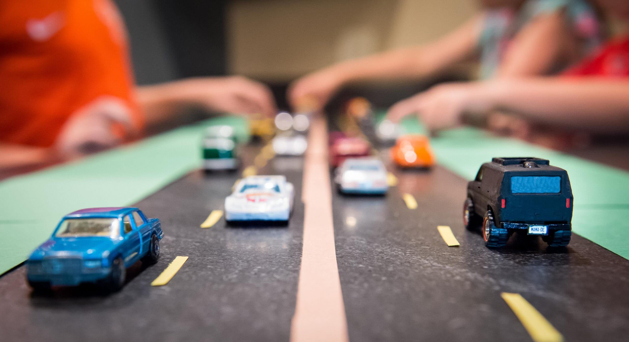 Toy cars are on a road mat. People stand over the road and reach toward the cars.