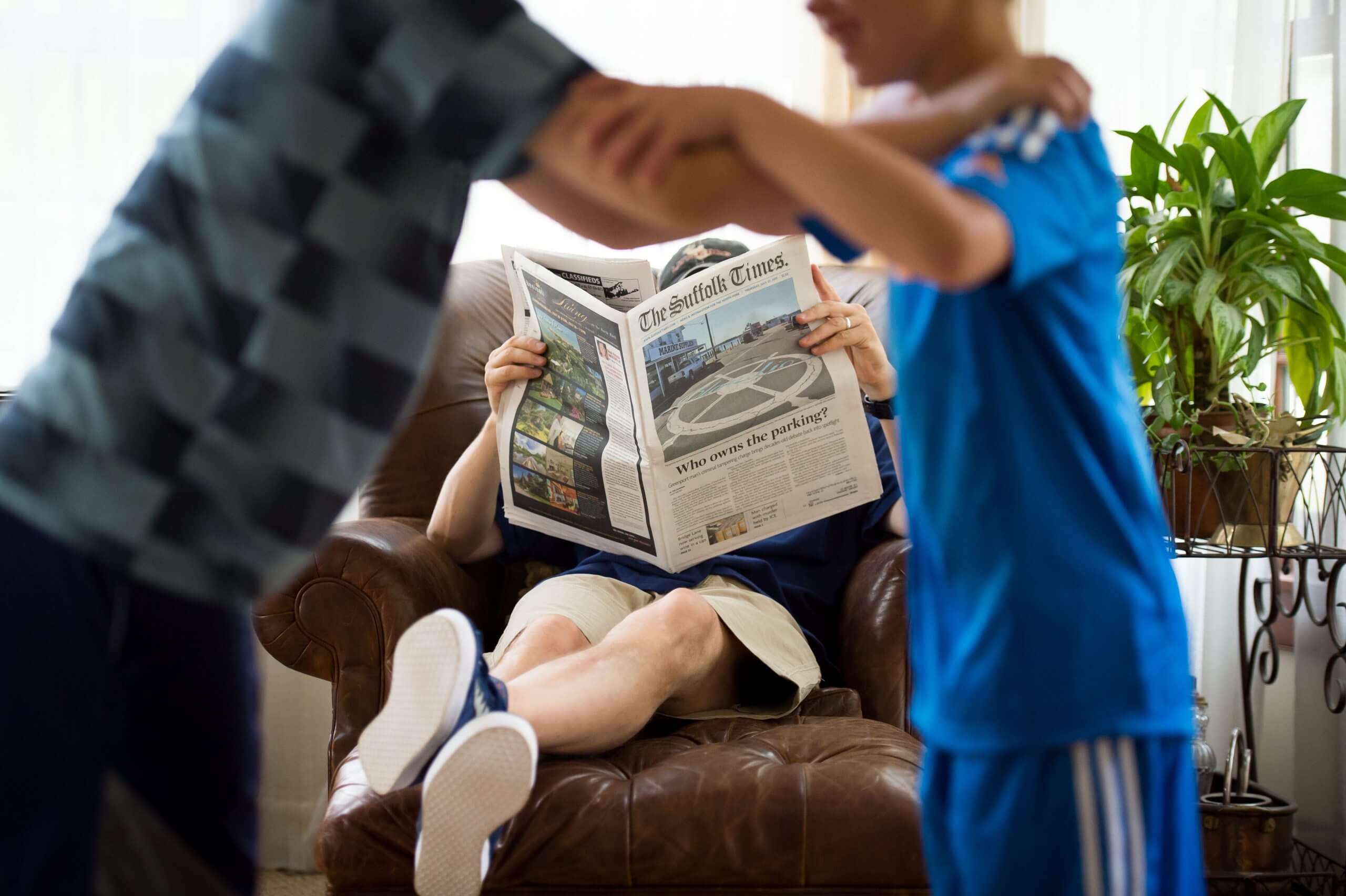 A focused view of a seated man with his feet up reading the newspaper. In front of the man, unfocused view, are two children leaning forward grappling each other.