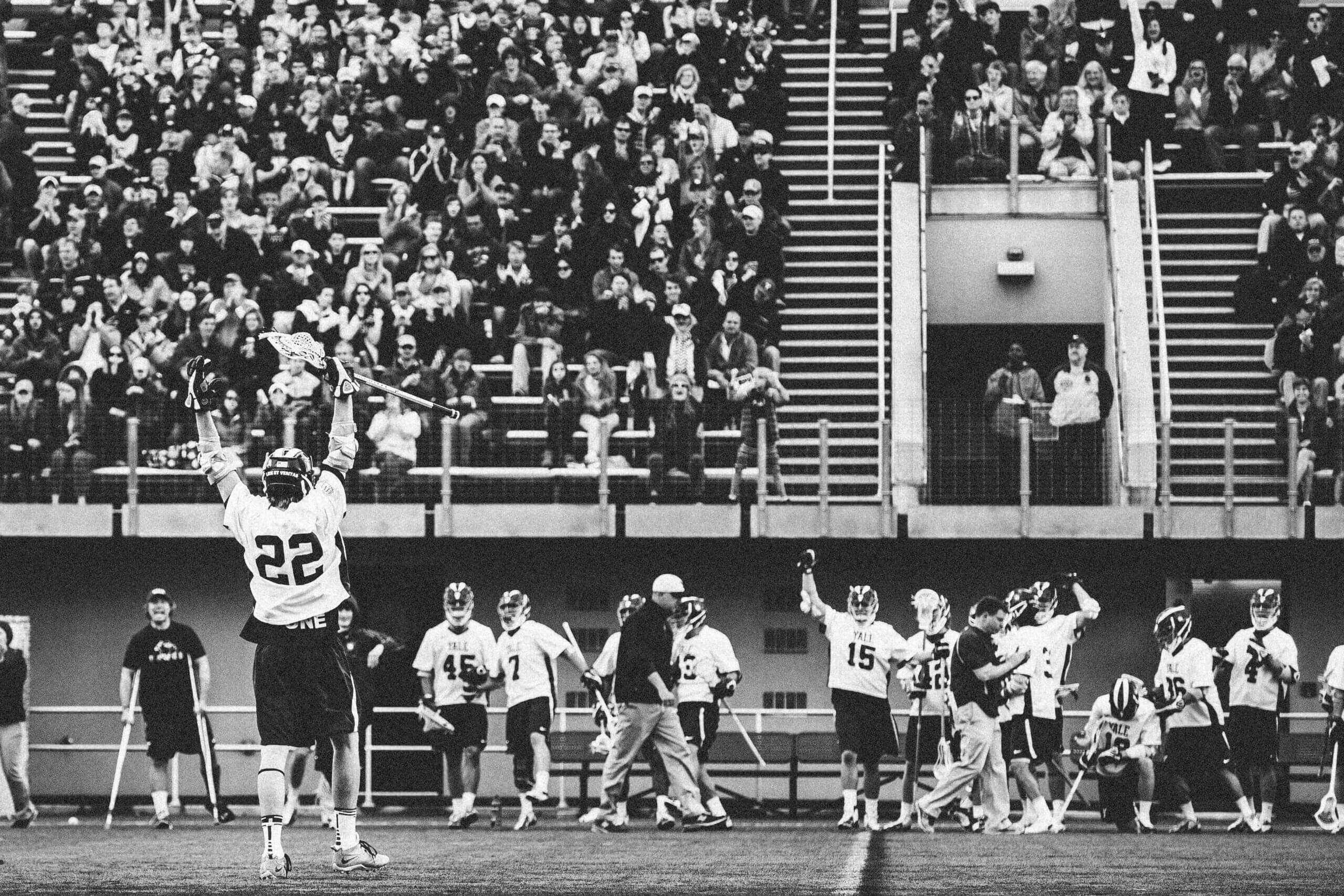 Black and white image of a lacrosse player celebrating a goal with the fans in the background.