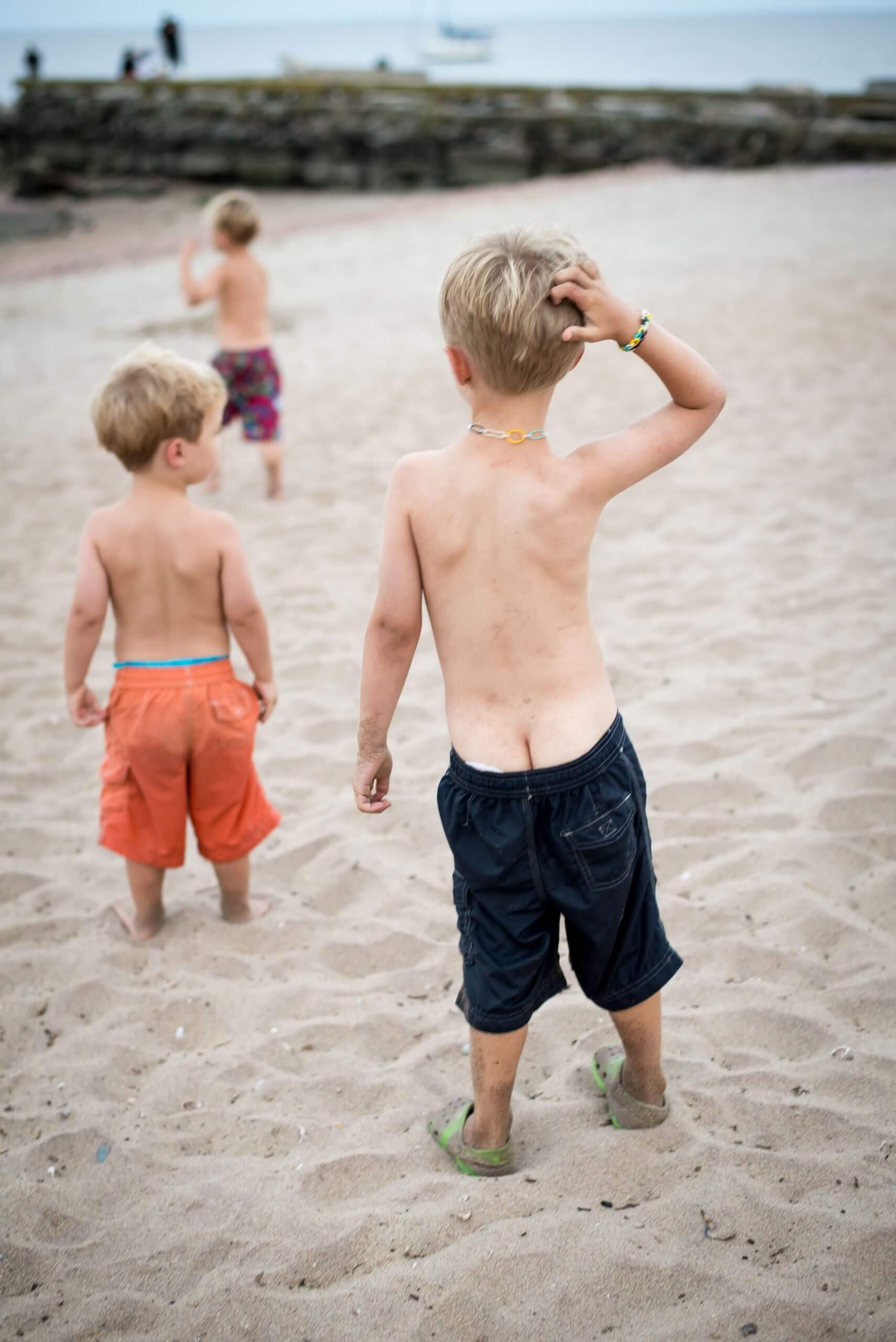 Three boys on a beach. One boy, his shorts falling and exposing part of his bottom, scratches his head and looks forward toward the other boys.