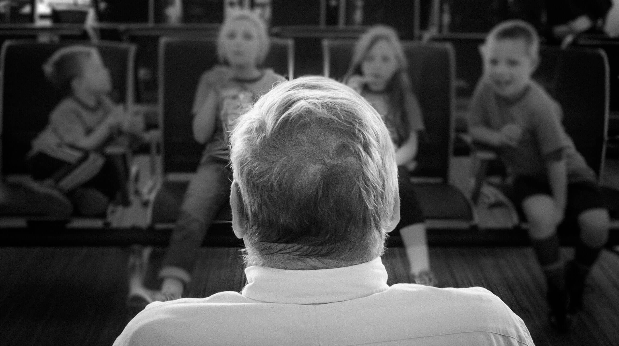 A focused view of the back of a man's head. The man looks forward to an unfocused view of four children seated in a theater.