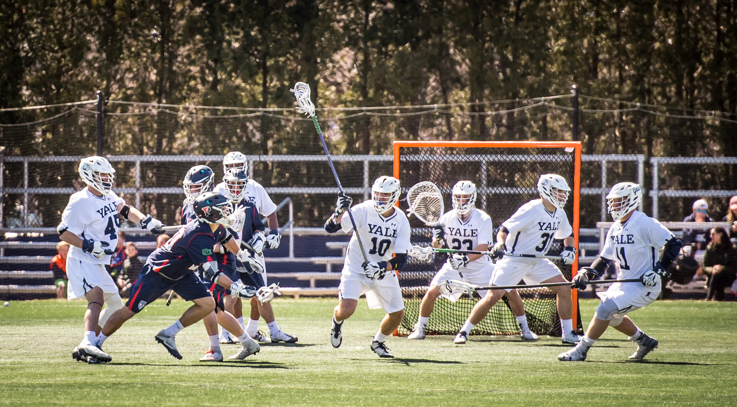 Lacrosse players defend the goal while an opponent runs toward them.