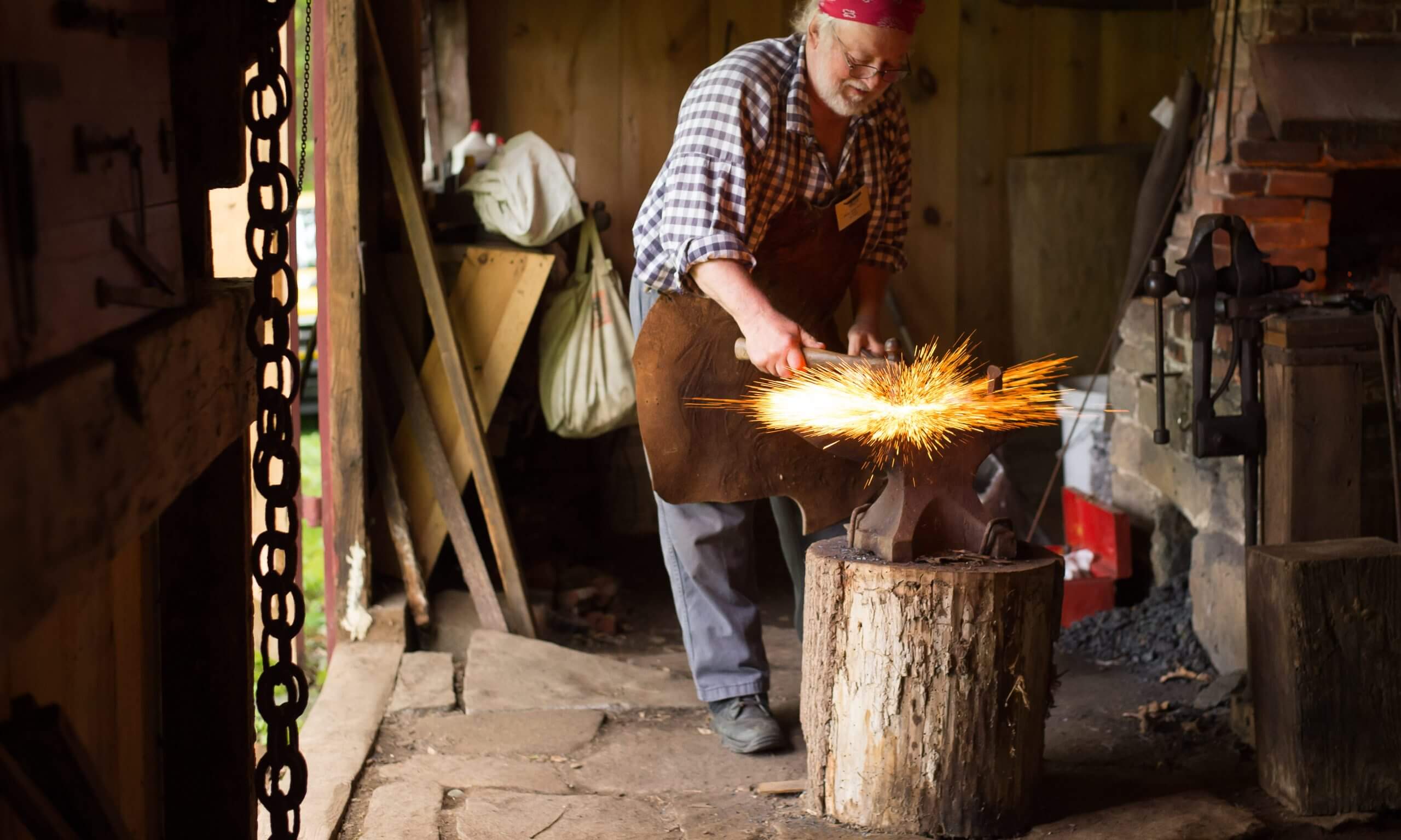 A metal smith wearing a bandana and a leather apron looks down at his work with tools in hand. Sparks fly from the item below him.