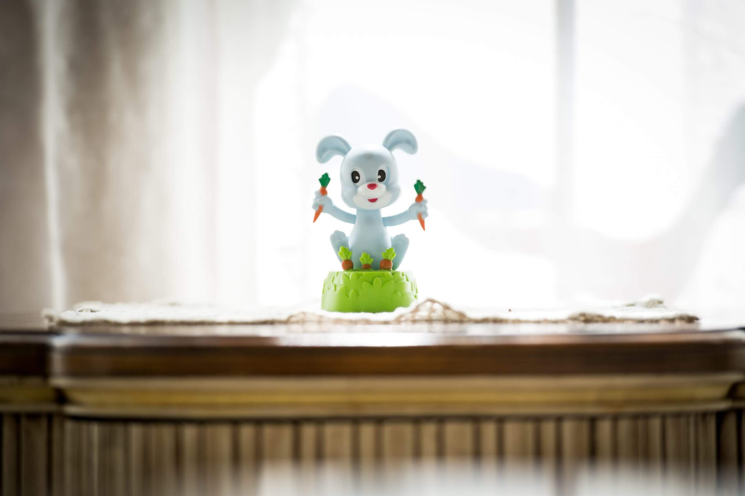 A smiling toy rabbit sits on a window sill. The rabbit moves from side to side, a carrot held out in both paws.