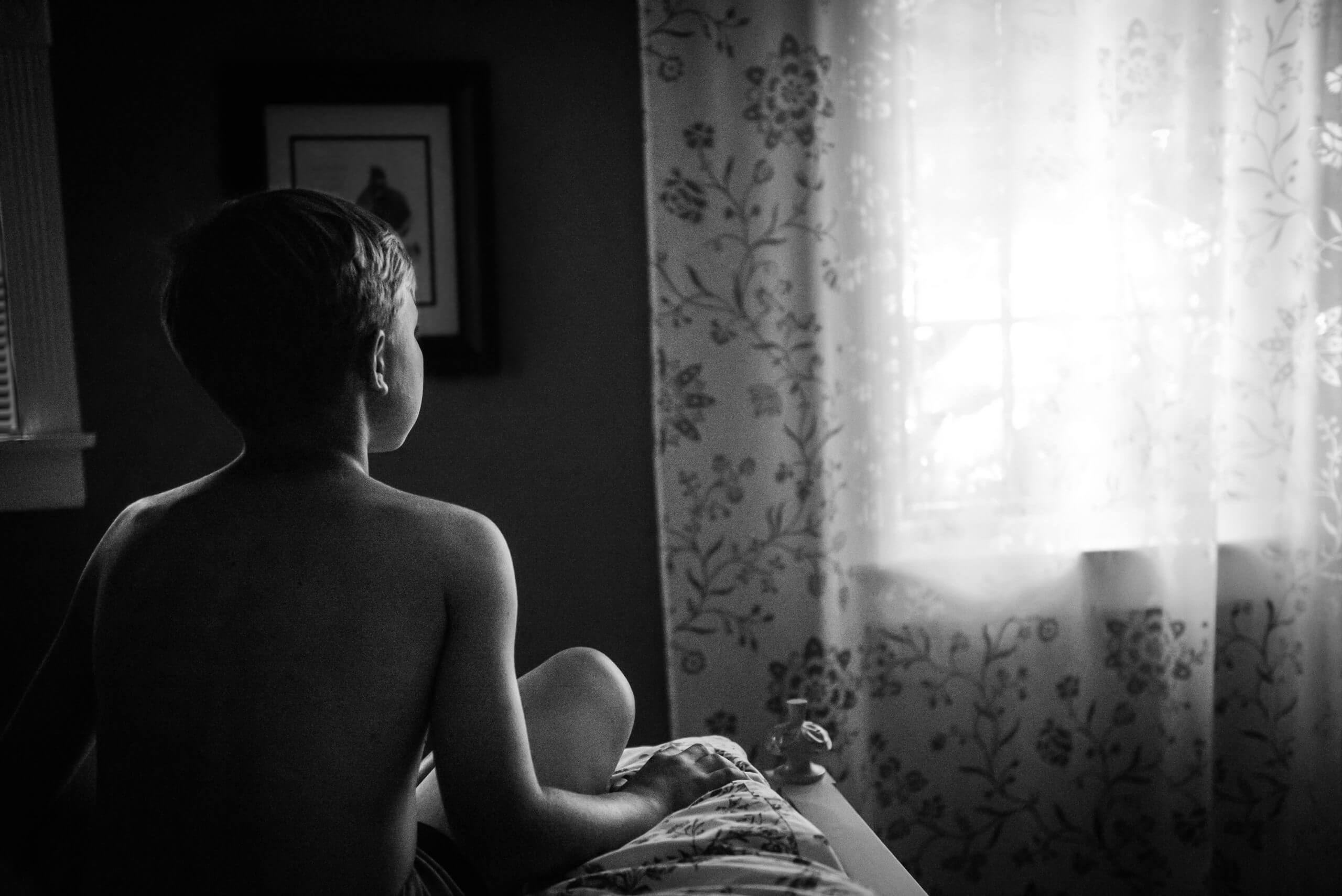 A boy sits on the foot of a bed and looks toward the curtained window in front of him. Light comes in through the window.