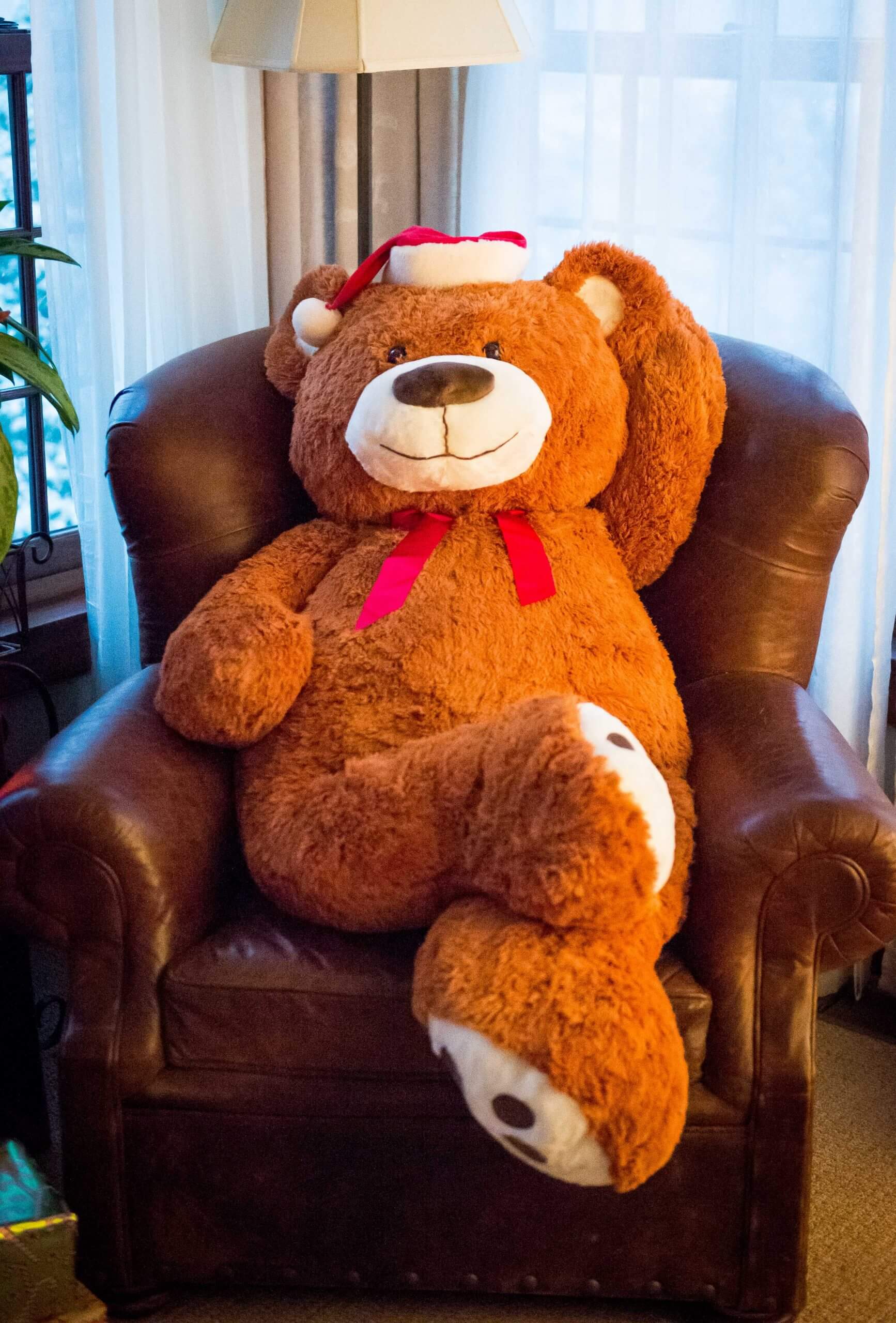 A large stuffed teddy bear reclines in a leather chair. The bear wears a santa hat.