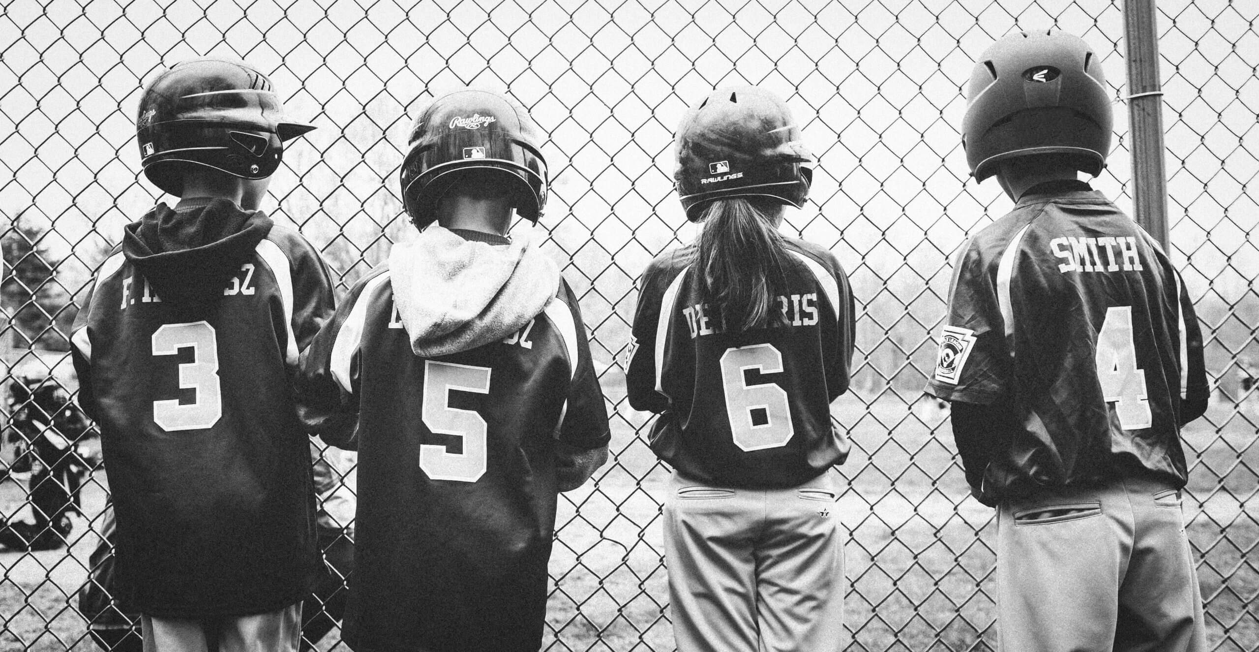 Four children in baseball uniforms stand behind a metal fence and look forward at the baseball field.