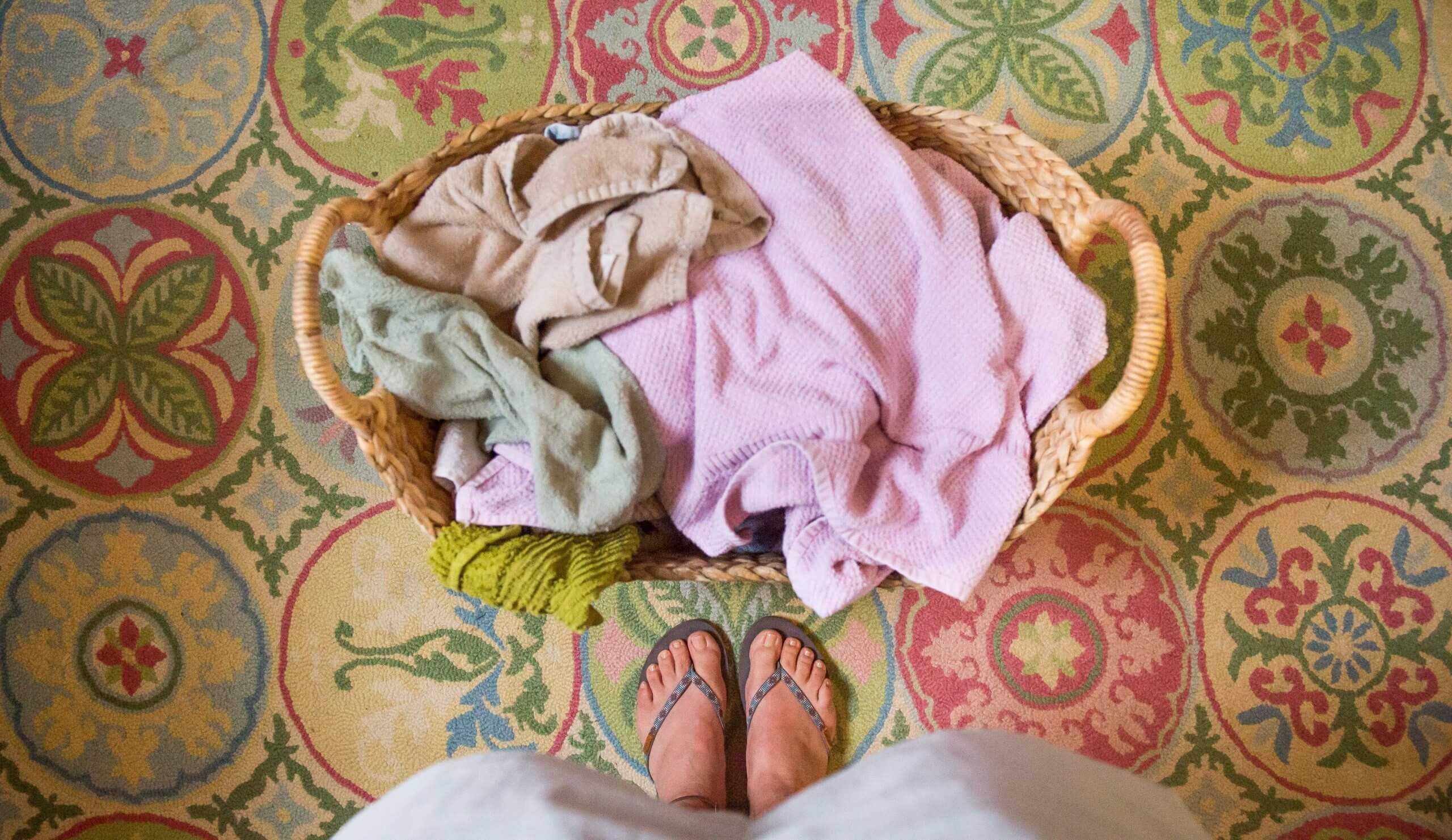 A basket of laundry is shown parallel to a person's feet. The person is standing beside the basket.