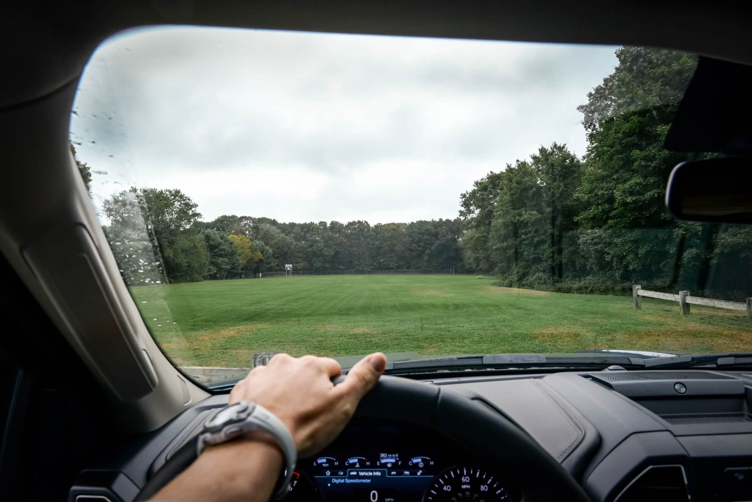 A view from the interior of a vehicle looking forward through the windshield from the driver's side. Outside is a field surrounded by trees.