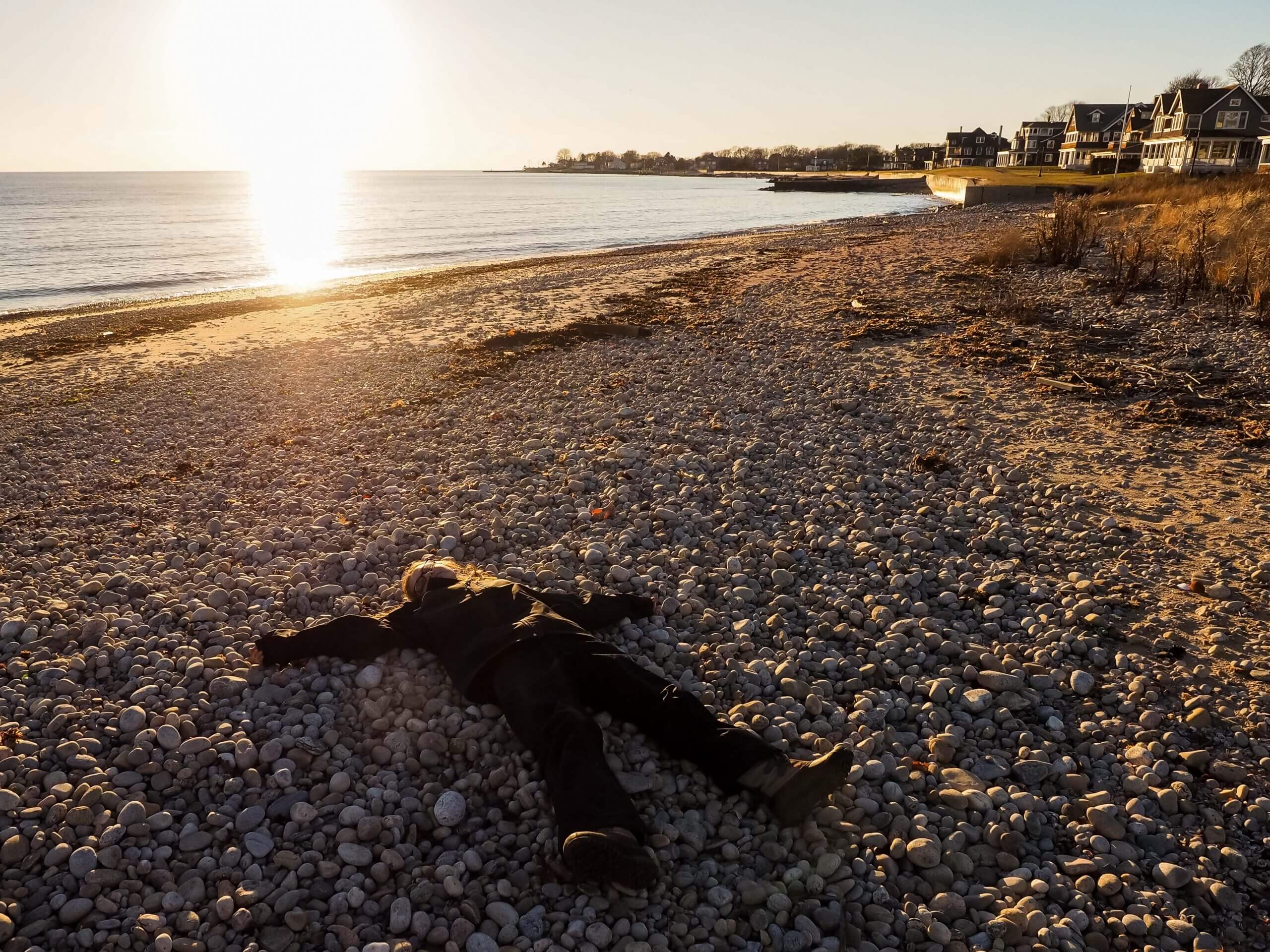A person lays outstretched on the pebbled surface of a beach. The sun shines brightly.