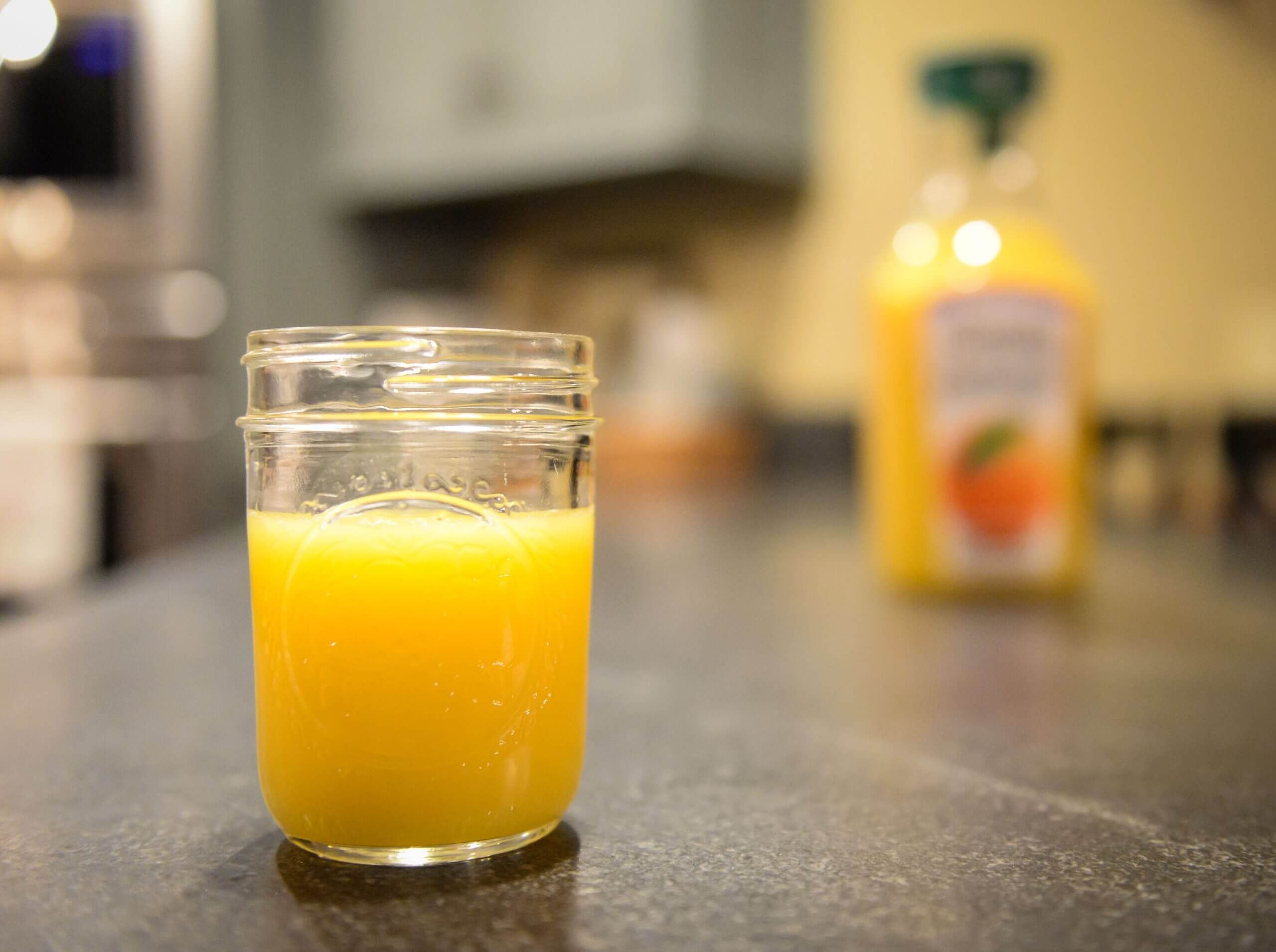A focused view of a glass of orange juice on a counter, in front of an unfocused view of the container of orange juice.
