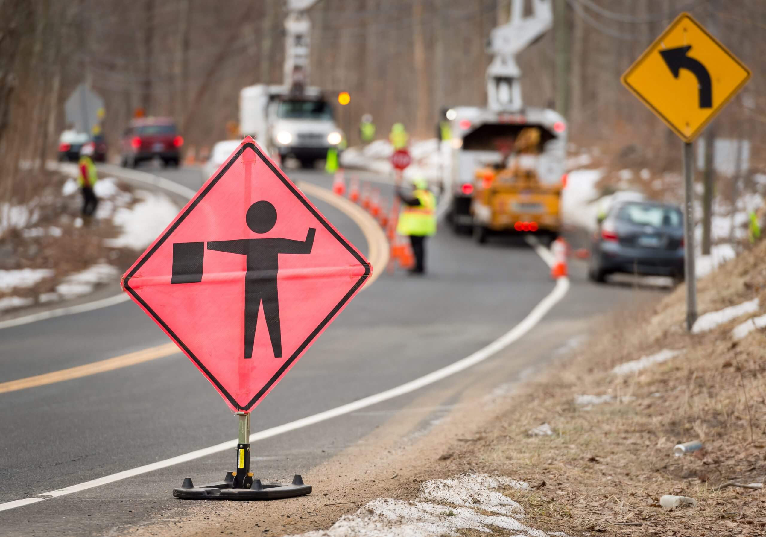 A focused view of a flagger ahead sign is shown on the side of the road. Behind the sign is an unfocused view of a traffic flagger, traffic cones, and two lift trucks.