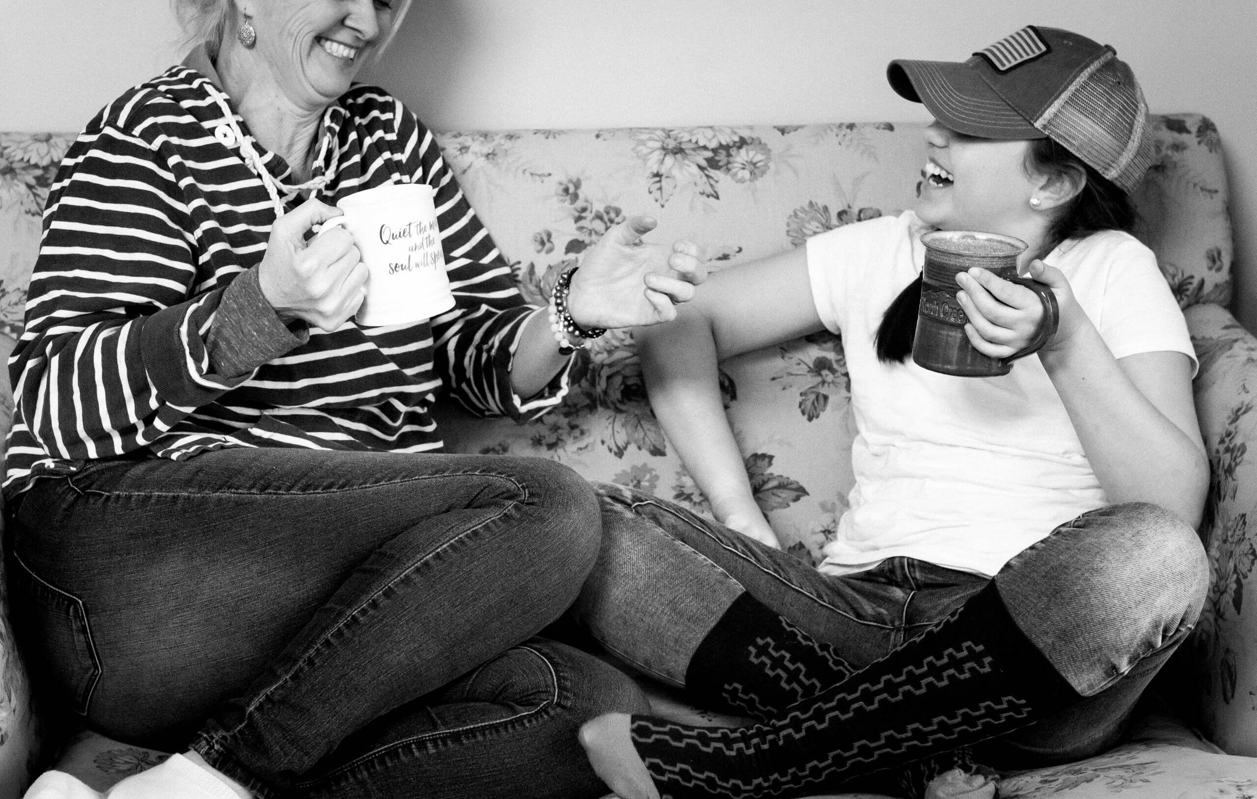 A woman and a child sit on a couch holding mugs and laughing.