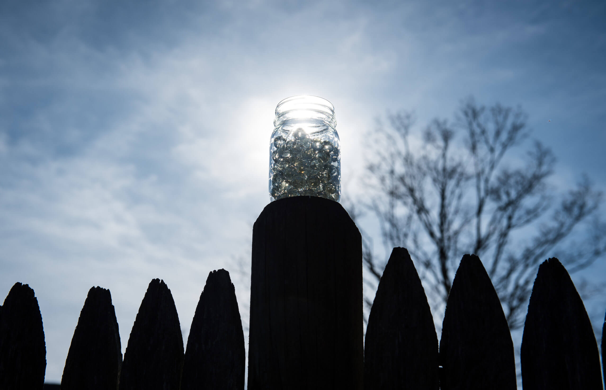 A jar of clear marbles is on top of a fence post. The sun shines through the marbles.