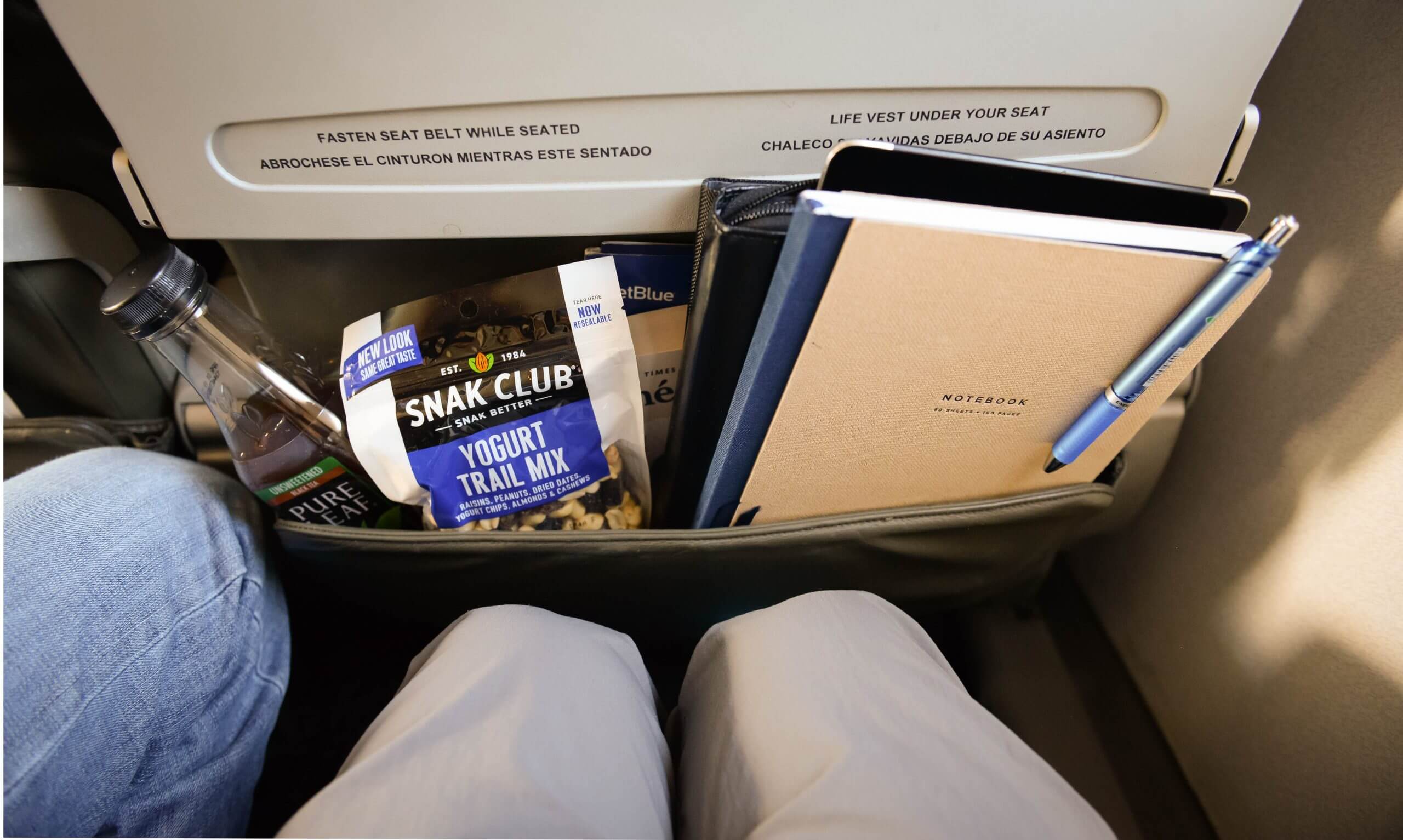 The back of an airplane seat includes a pocket with the following items: bottle of iced tea, bag of trail mix, notebook and pen.