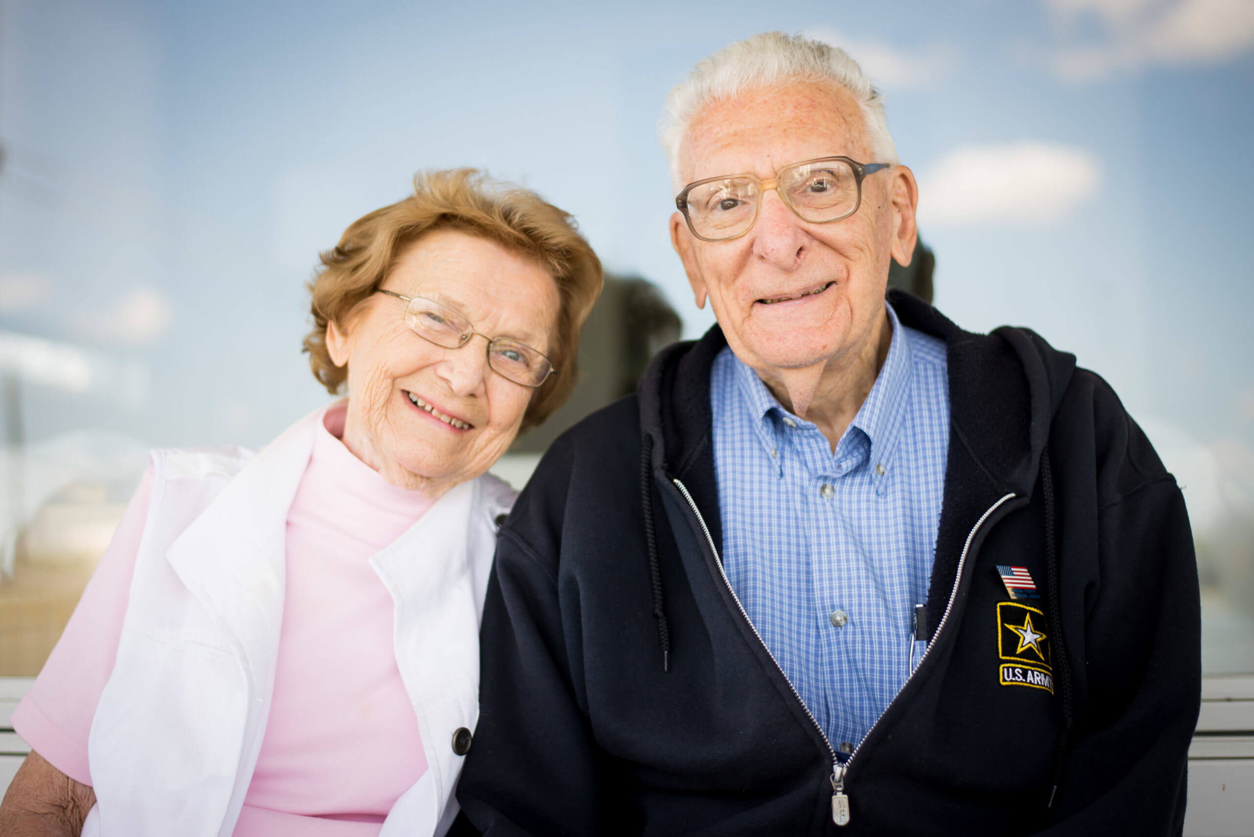 A portrait of an older couple smiling. The woman leans toward the man. The man wears a sweatshirt with a US Army patch.