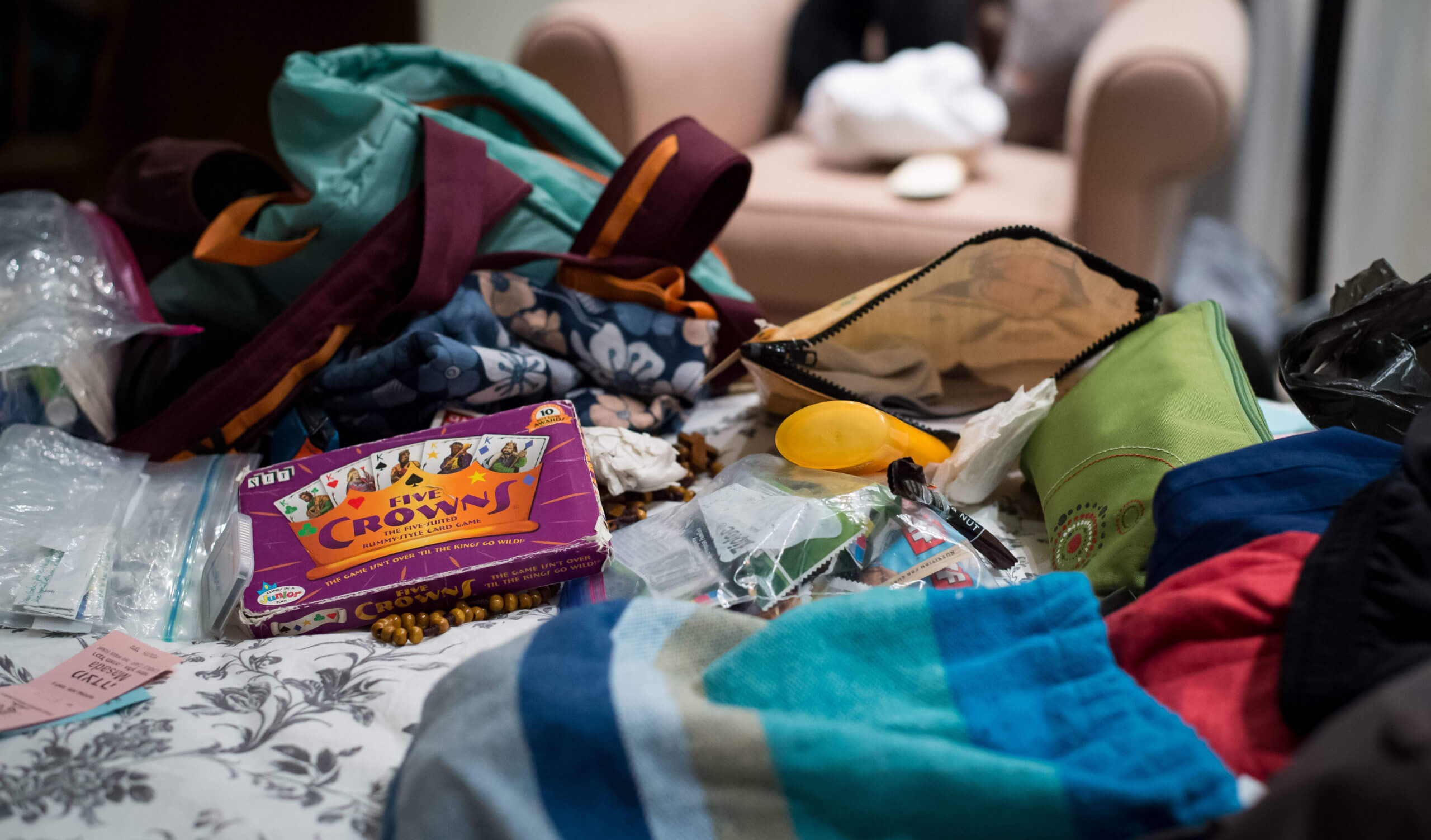 Items cover the surface of a bed and include backpacks, plastic bags, zippered pouches, and a card game labeled "The Crowns."