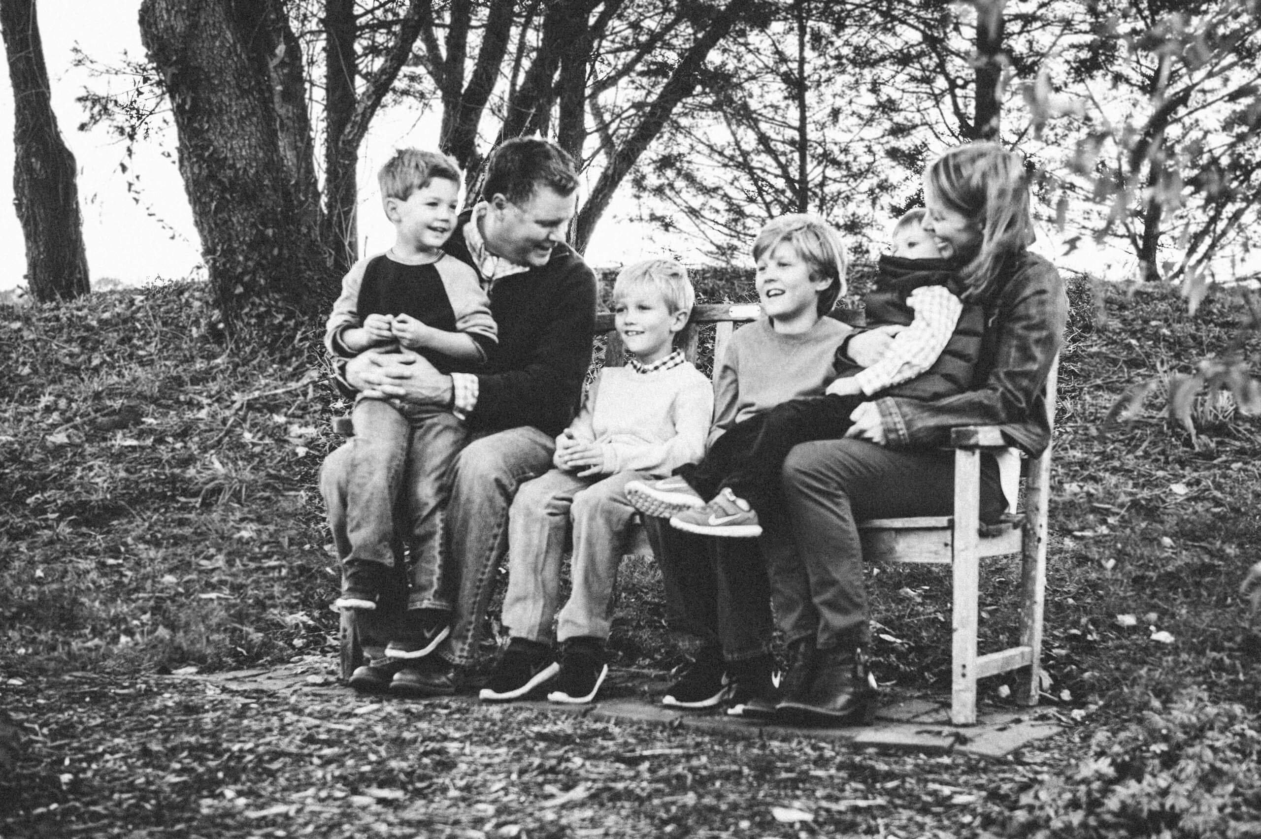 A portrait of a family sitting on a wooden bench outside. A man and woman sit on either side of the bench and smile down at the four children sitting between them.