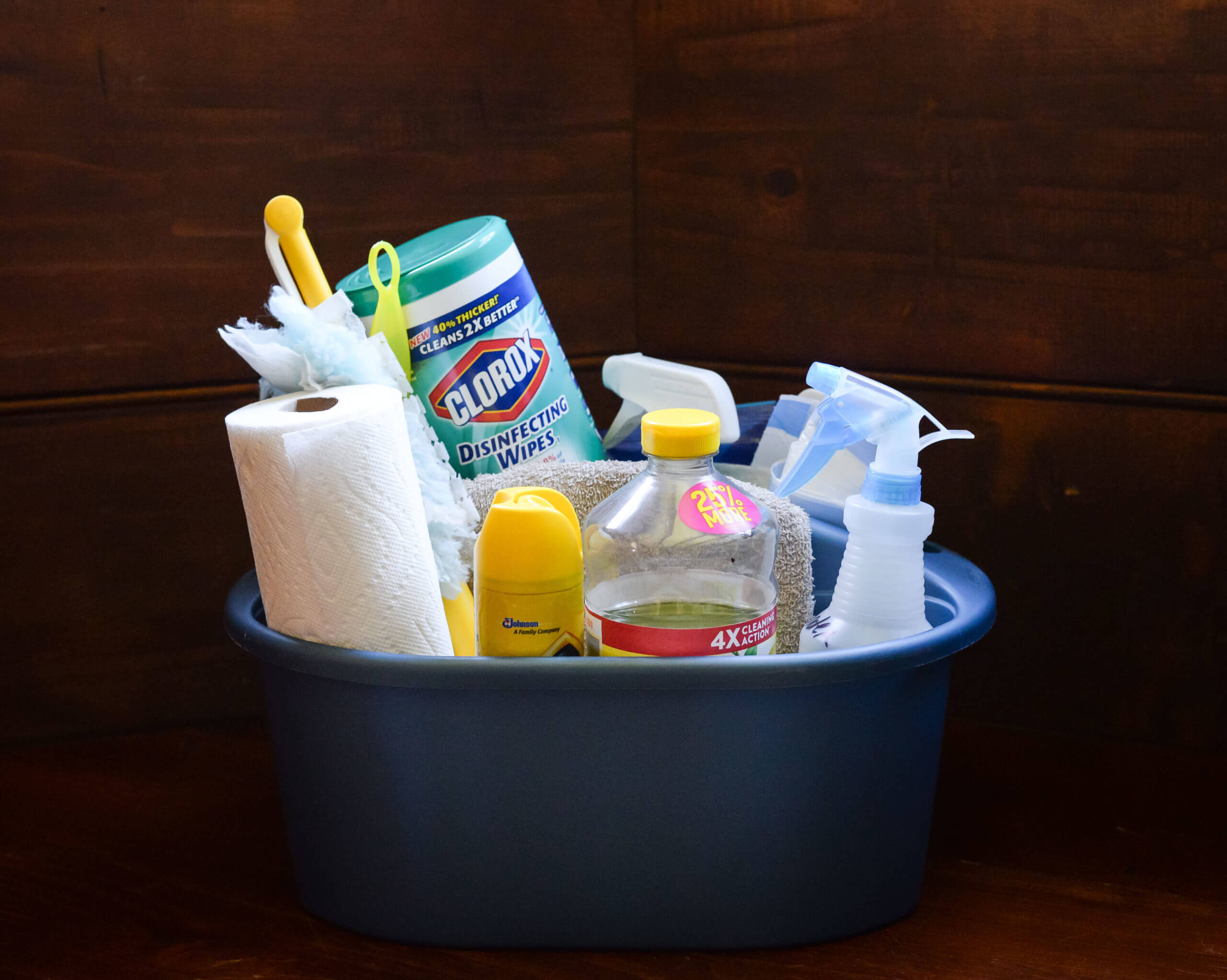 A plastic tub is filled with various cleaning supplies including Clorox wipes, paper towels, a duster, spray bottles, and rags.