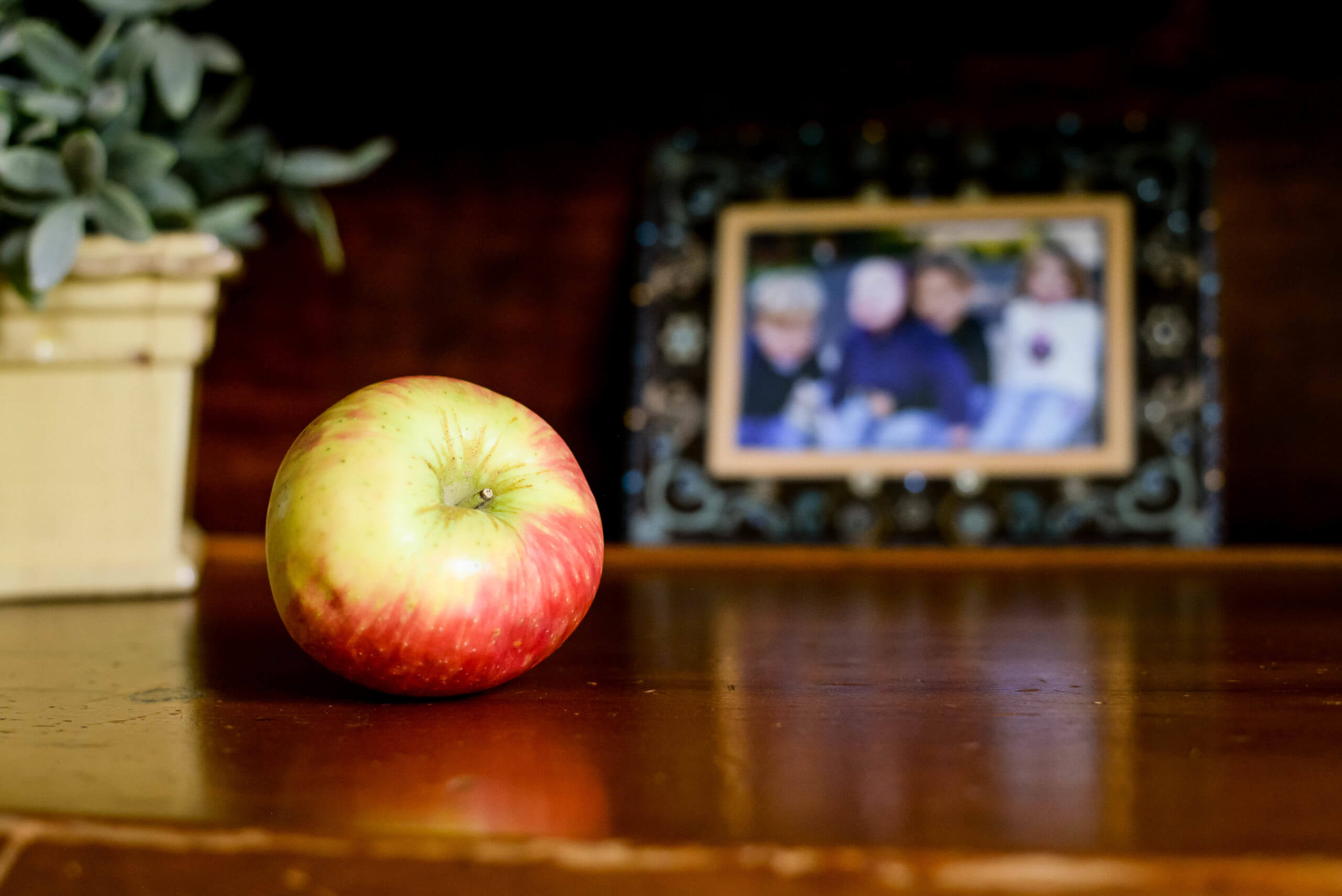 A focused view of an apple on a table. Behind the apple is an unfocused view of a framed family portrait.