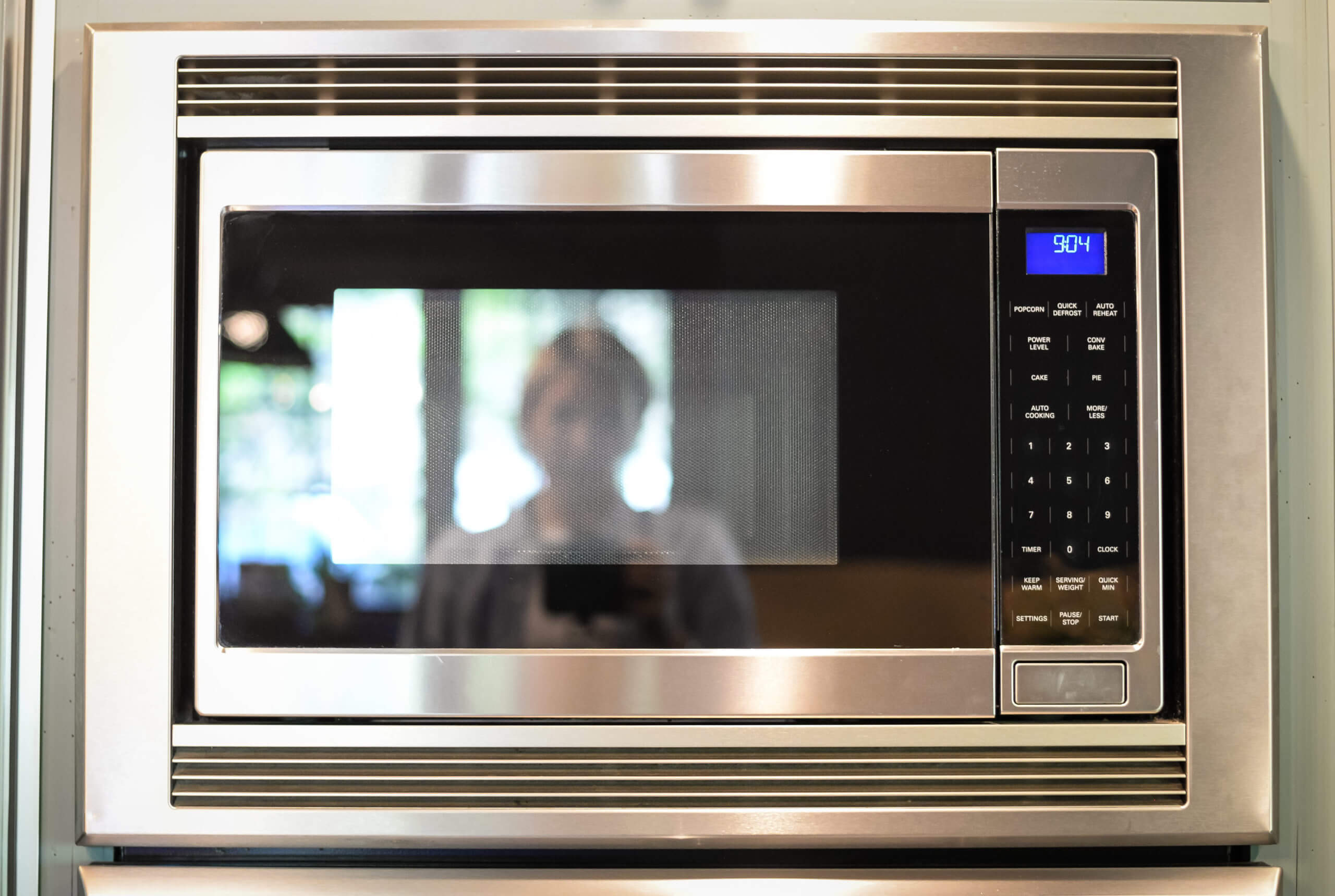 A stainless steel microwave . The reflection of the photographer is shown in the glass of the microwave.