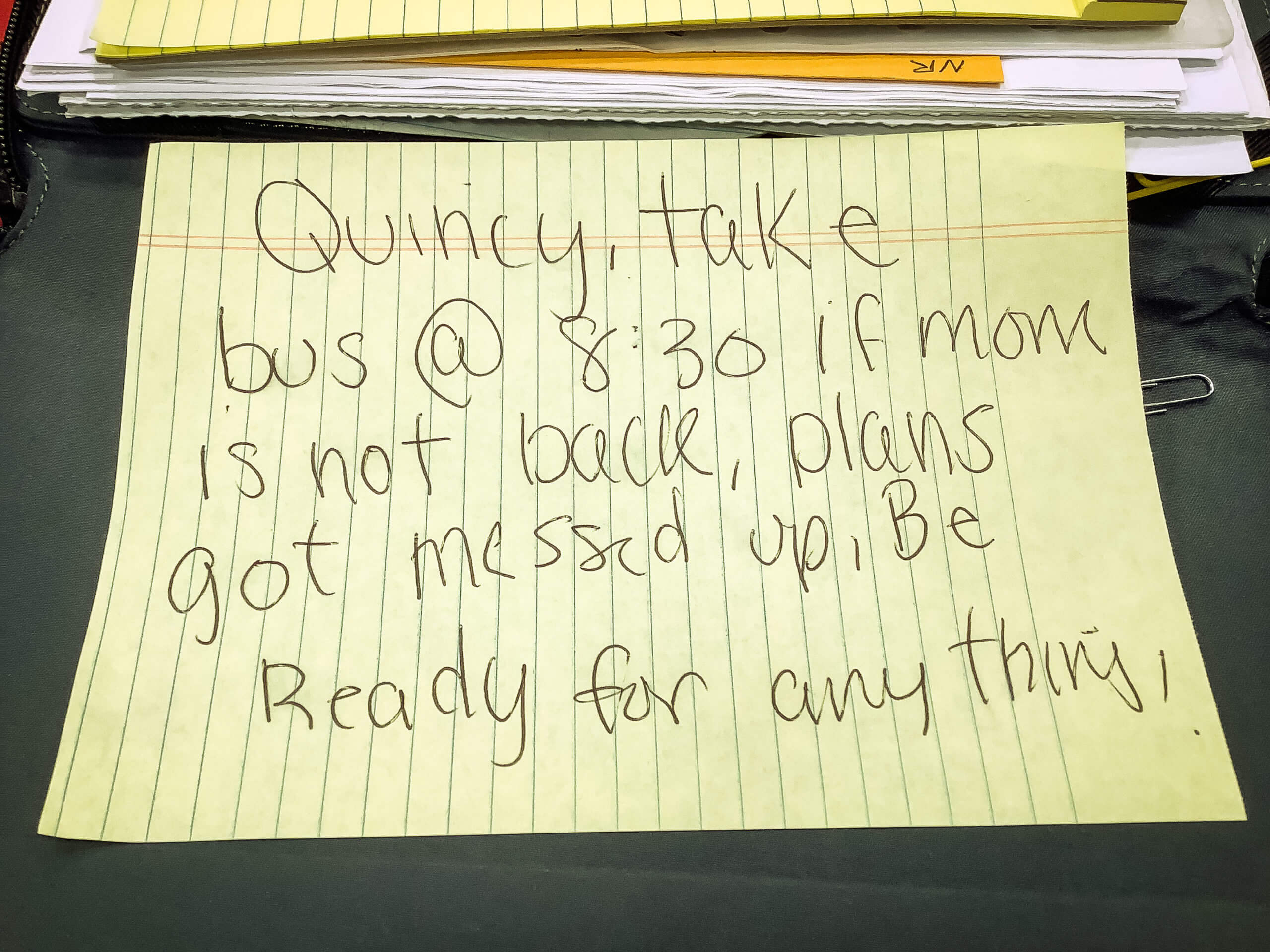 A piece of paper with a handwritten note. The note reads "Quincy, take bus @ 8:30 if mom is not back, plans got messed up, Be Ready for anything!"