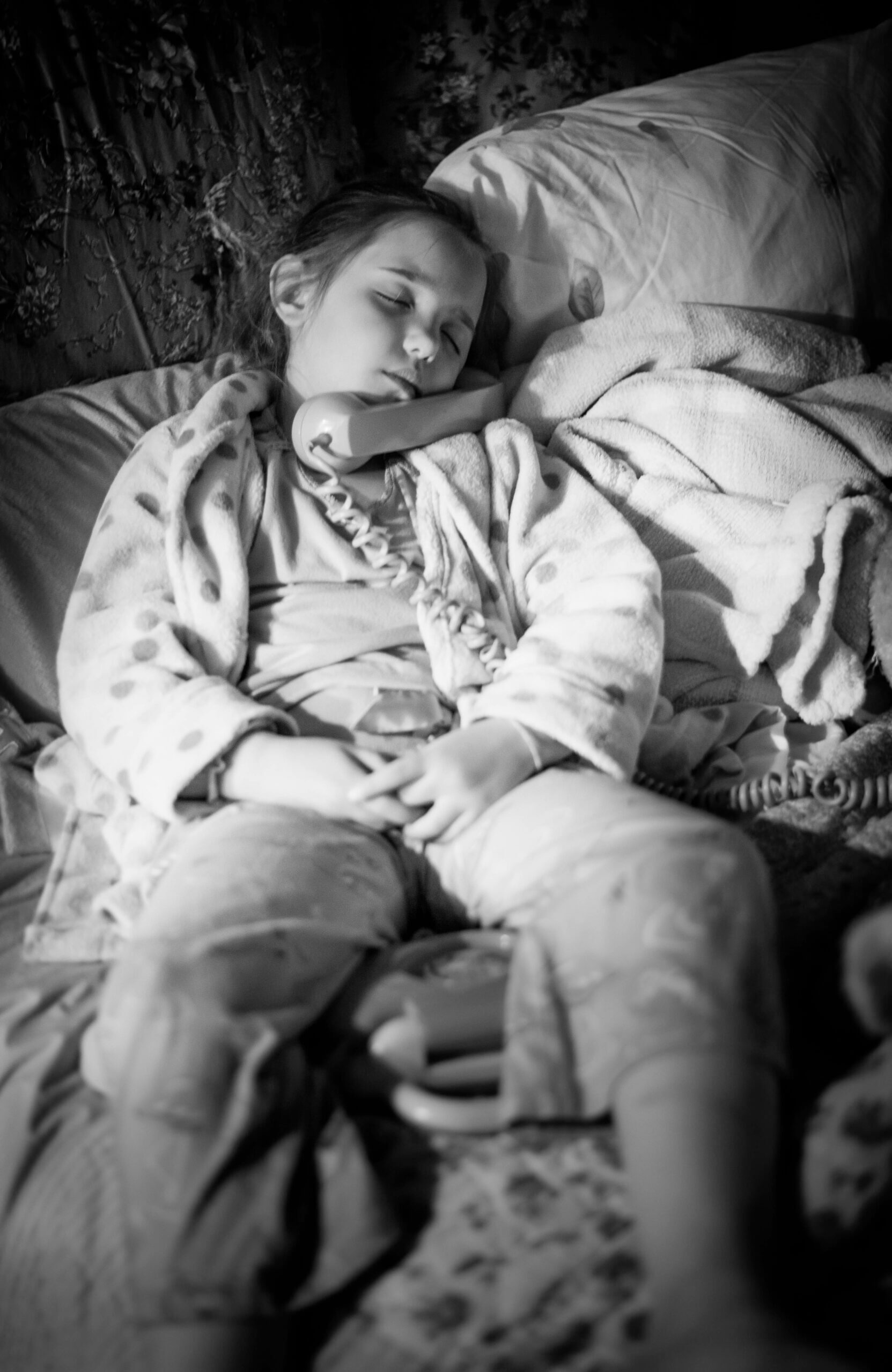 Black and white photo of a little white girl with a phone at her ear while she's sleeping.