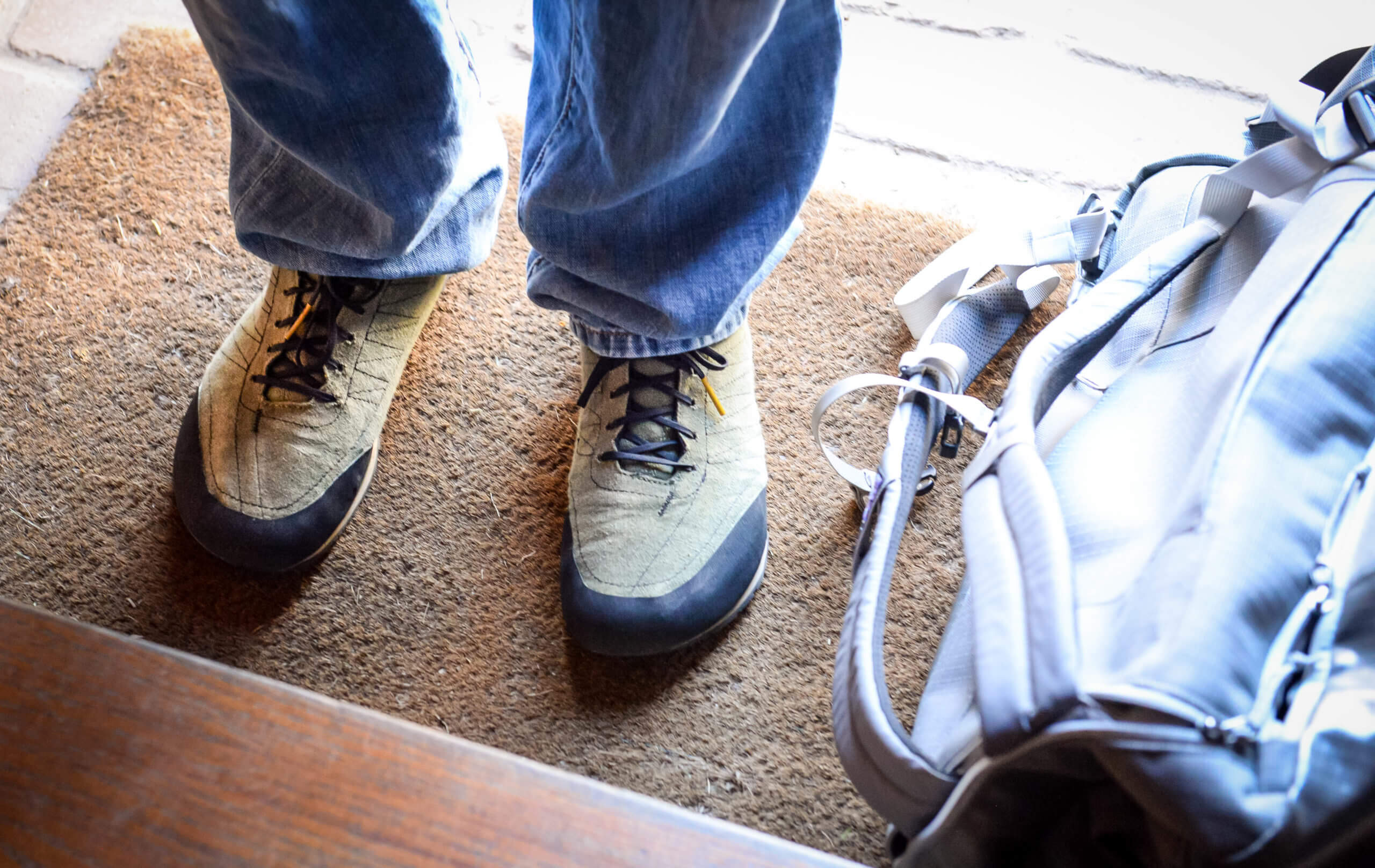 A view looking down at a person's shoes on the mat of a front doorstep. A backpack is on the mat.