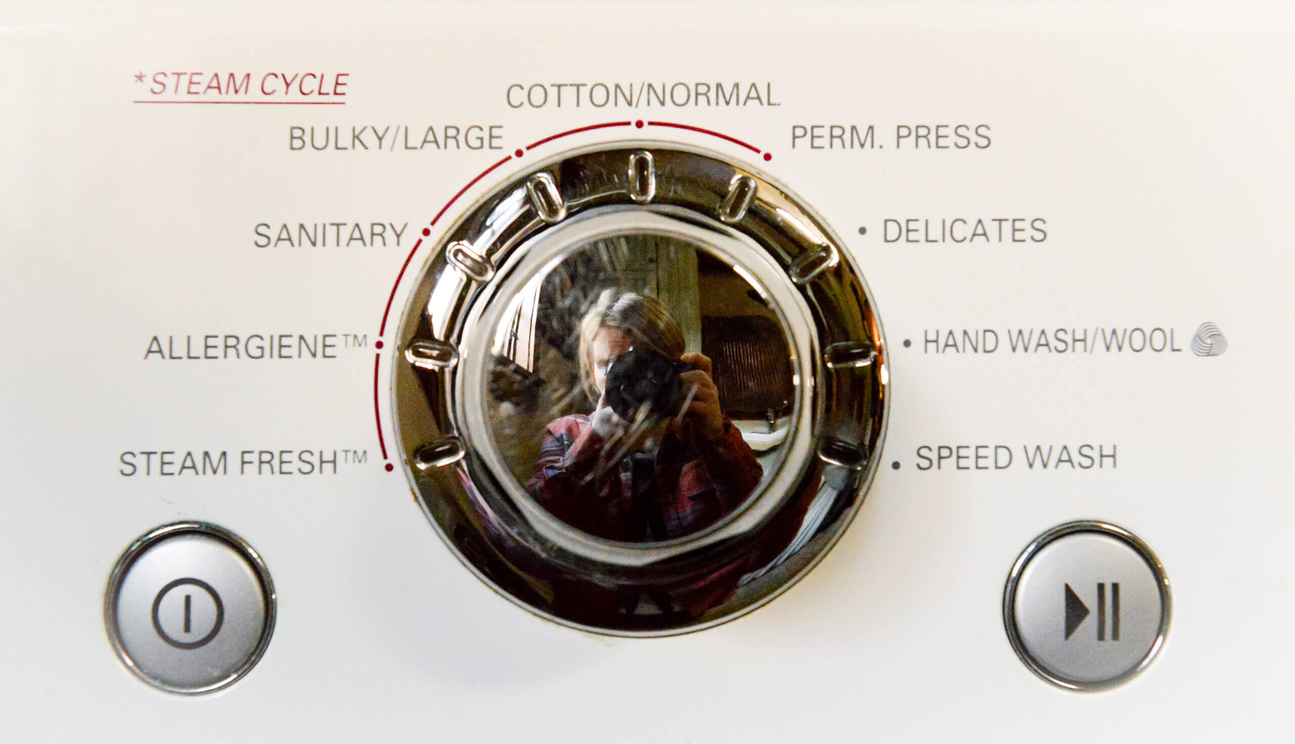 The settings dial of a washing machine. The dial is chrome and the photographer's reflection is shown at the center of the dial.