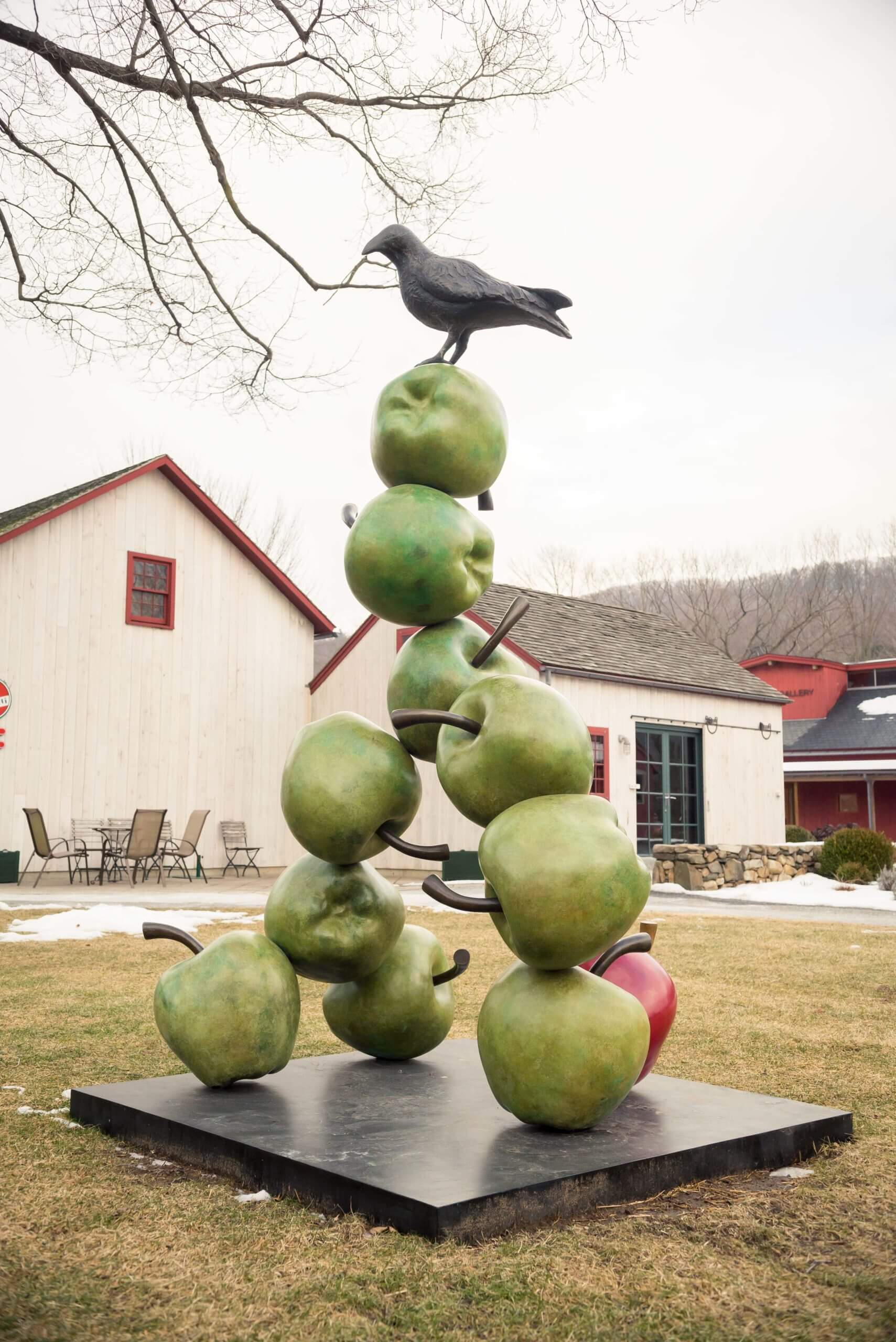 A statue of a blackbird on top of a narrow stack of apples outside on the lawn of a nearby building.