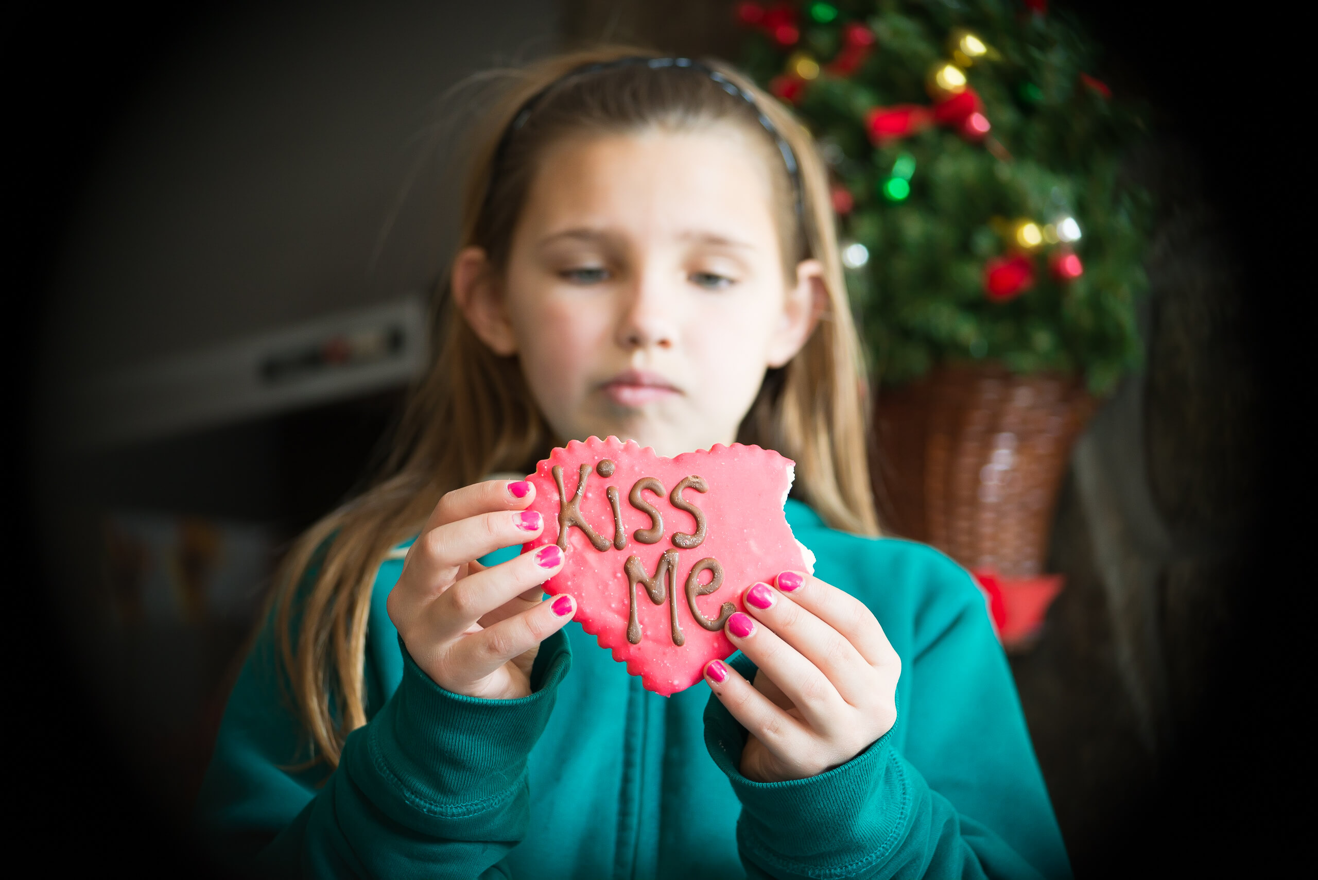 A cookie held forward by a child. The child looks down at the cookie. The cookie is a heart shape, has a bite taken out of it, and reads "kiss me."