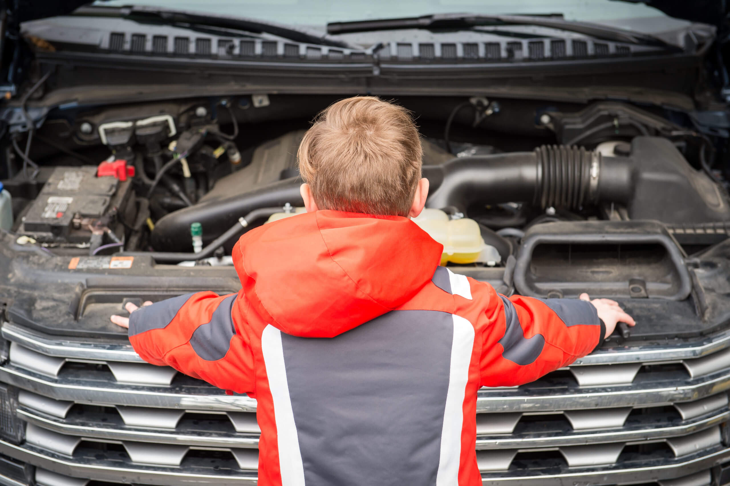 A boy stands looking down under the open hood of a vehicle.
