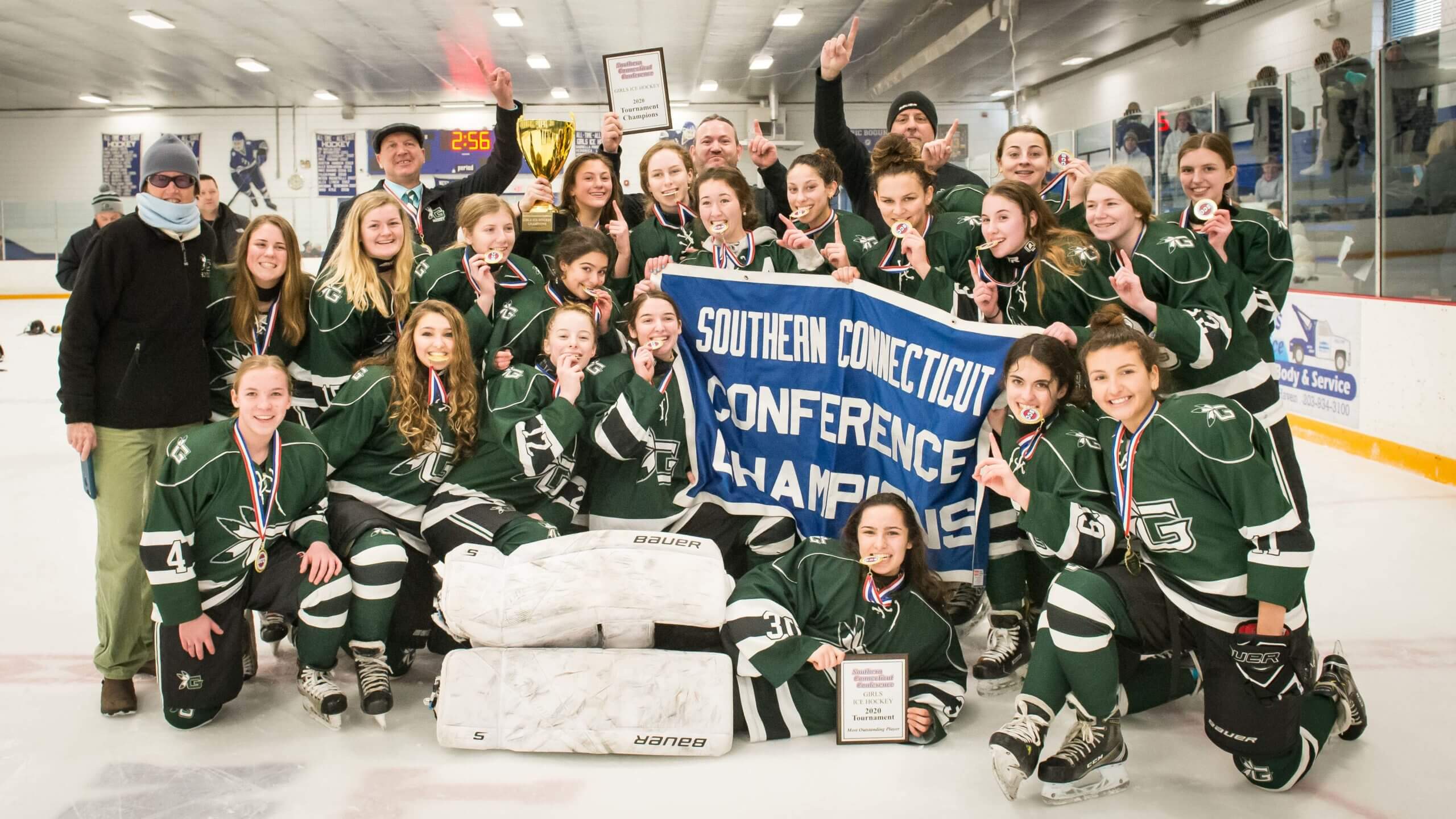Young female hockey players in uniform positioned in a group photo beside several adults in casual clothing. Everyone is smiling and a trophy is held up by a player. The girls hold up a sign that reads 