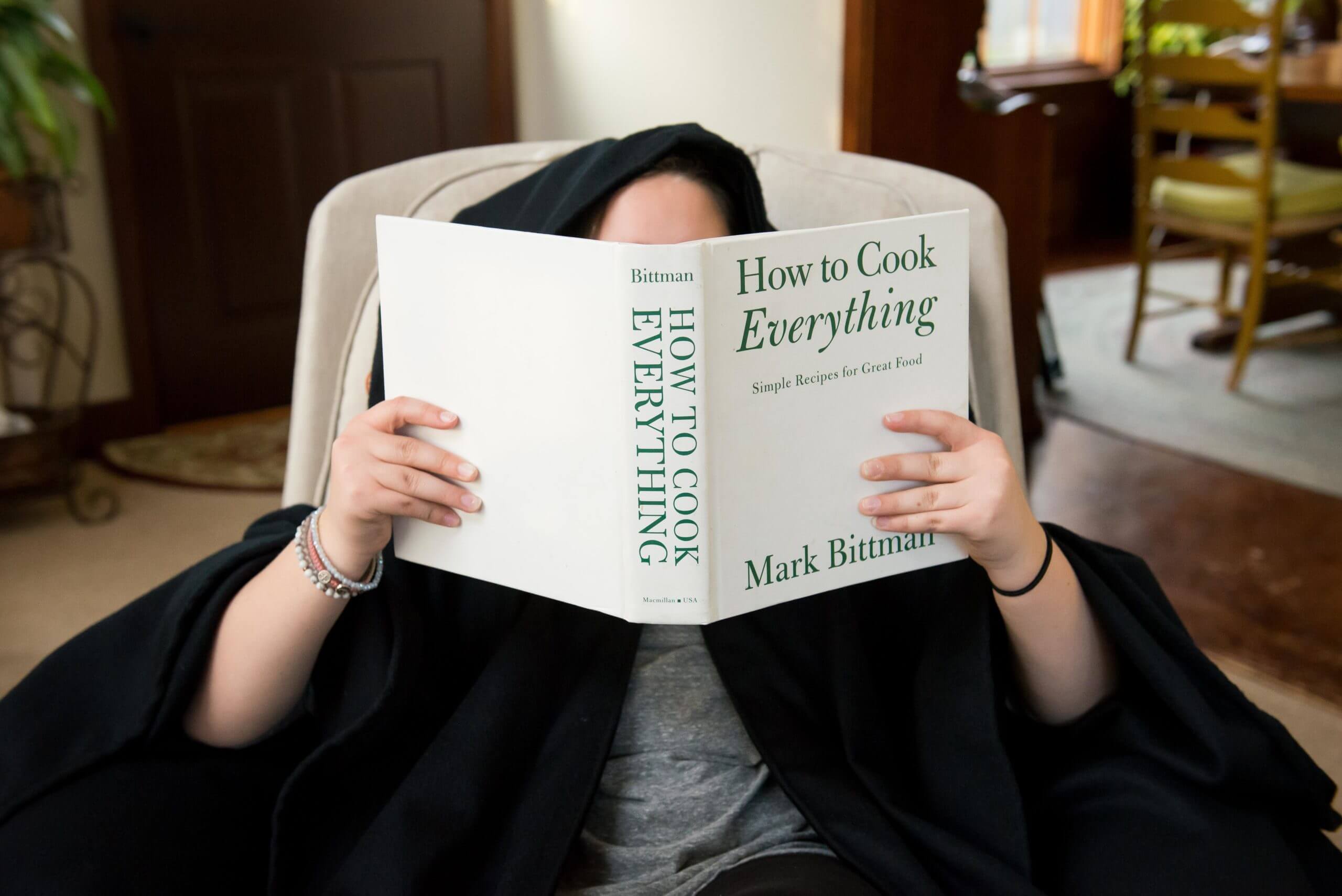 A person sitting in a chair is reading "How to Cook Everything" by Mark Bittman.