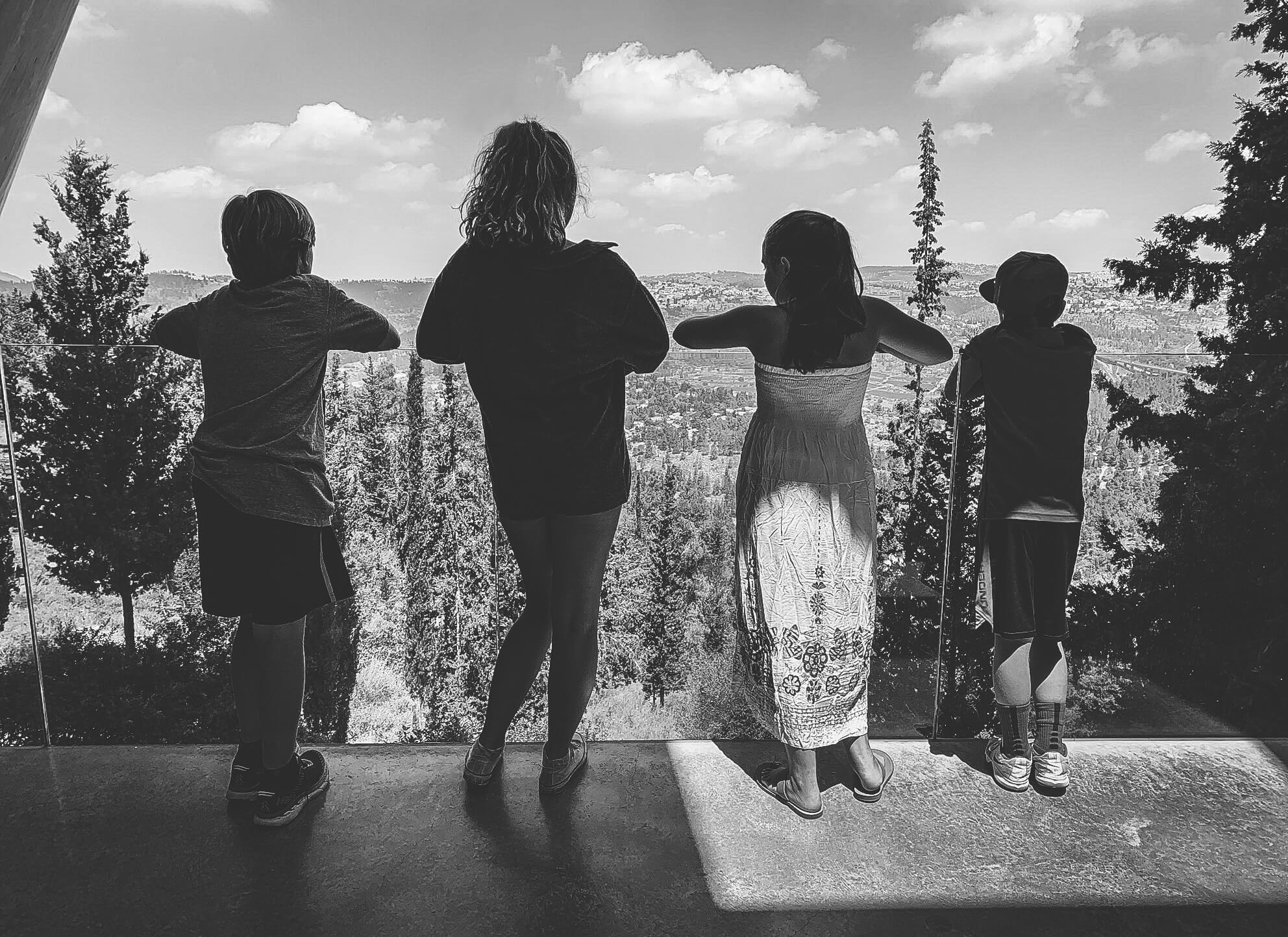 Four children stand outside against a plexiglass balcony shield. The children look down at the forest below.