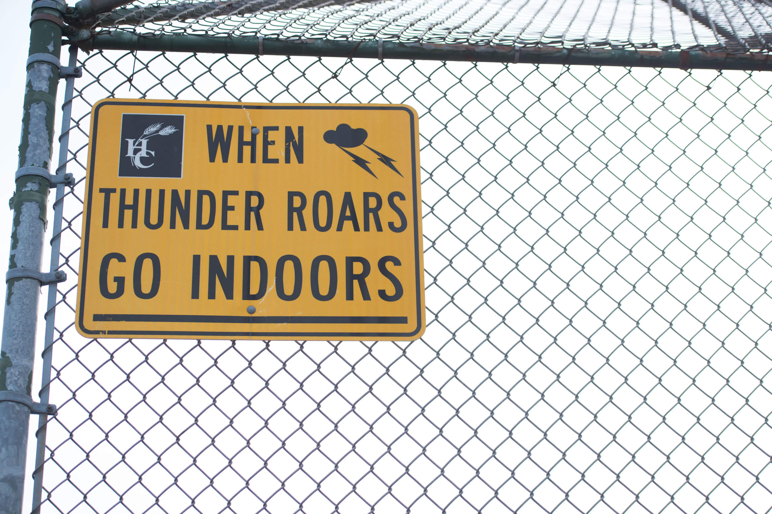 A sign attached to a metal fence reads, "When thunder roars go indoors." The word indoors is underlined and there is an outline of a cloud and lightning bolts.