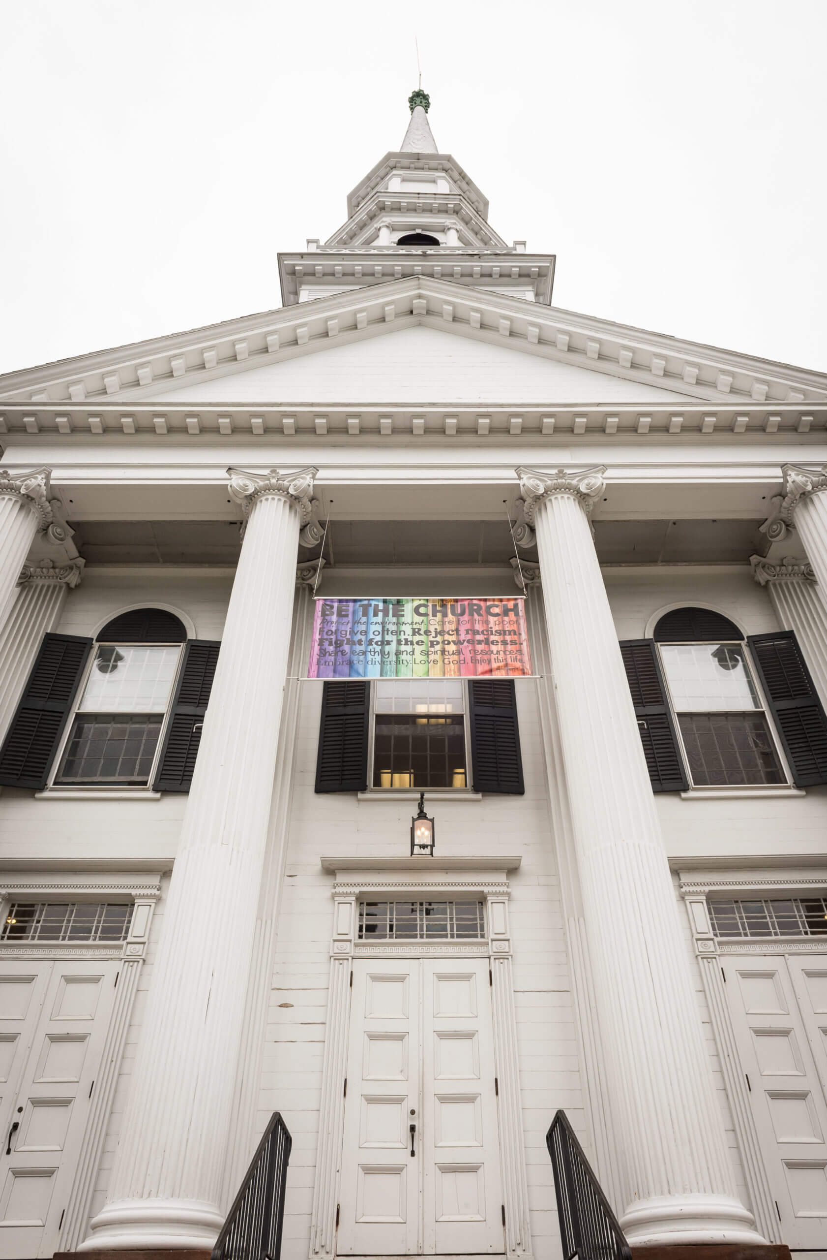 A view looking up at the front of a church. A sign hangs above the doors of the church. The sign is rainbow colored.