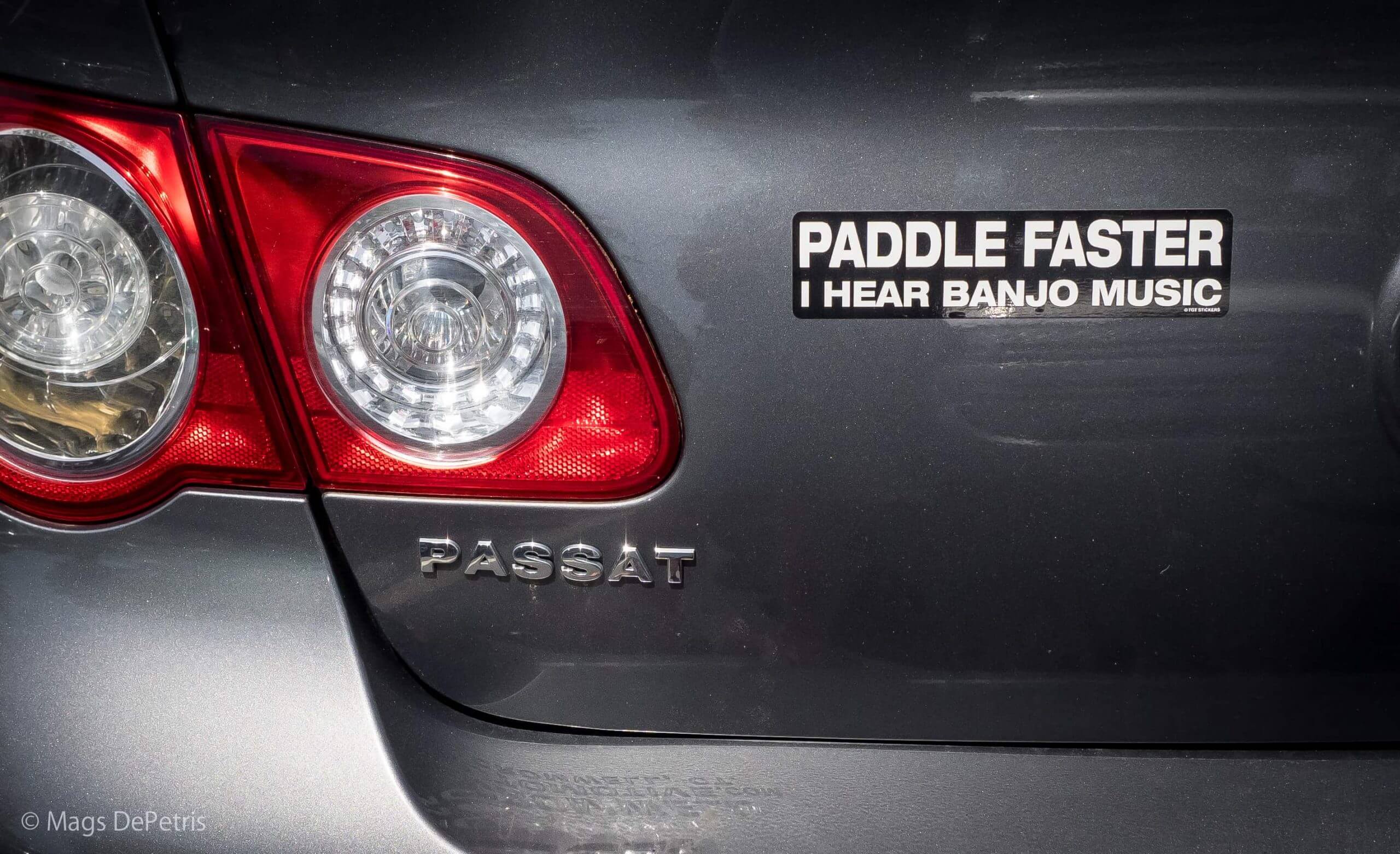 A tail light and bumper of a Passat car. A bumper sticker reads, "paddle faster I hear banjo music."