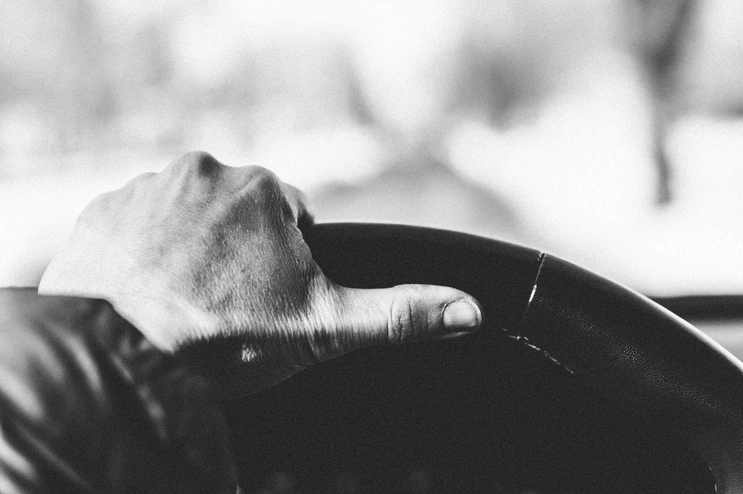 Woman's left hand tightly grasped on steering wheel, outside windshield out of focus, black and white photo.