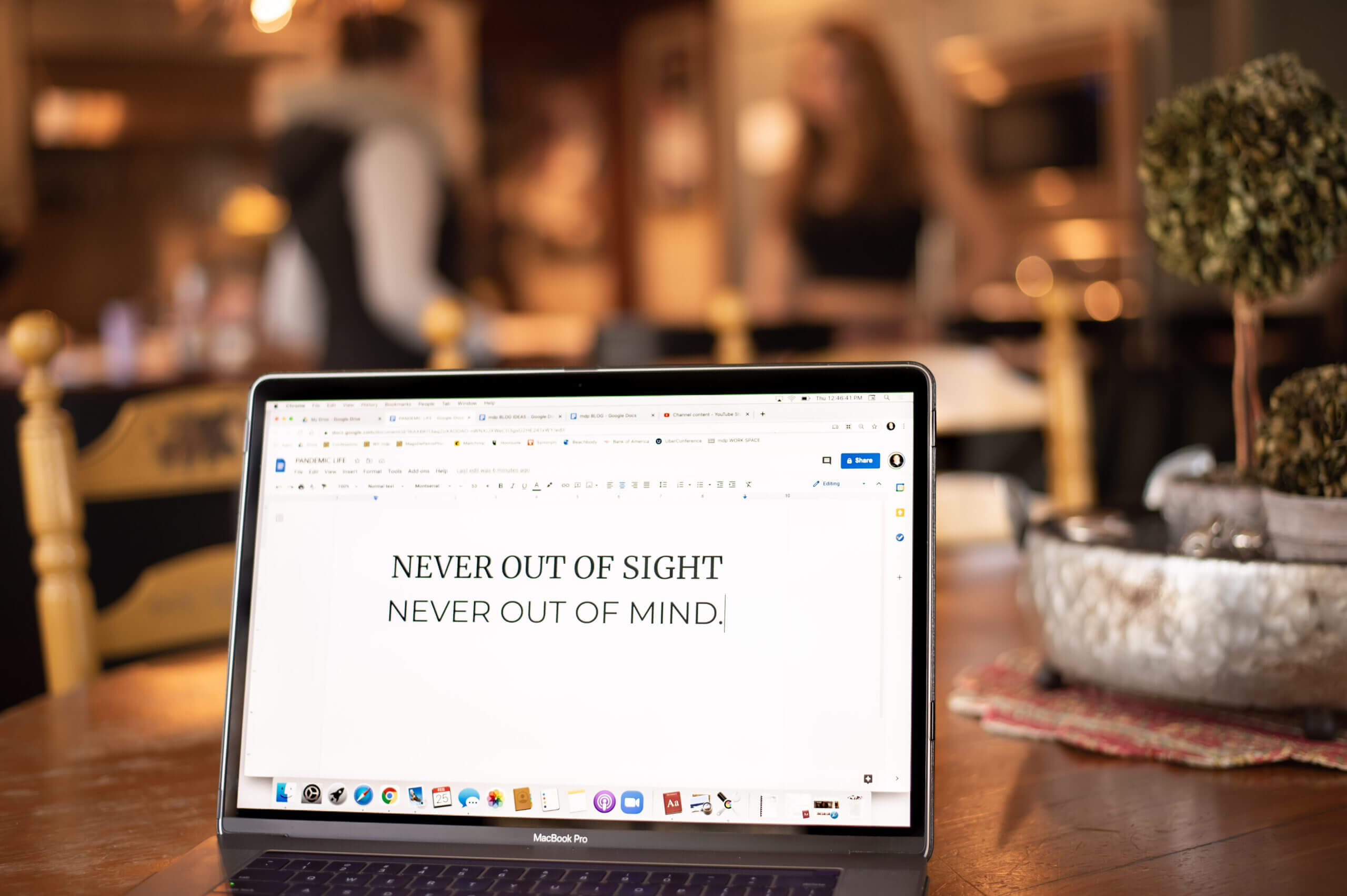 A MacBook Pro laptop sitting on a table with the phrase, “Never out of site, never out of mind” typed on the screen and the 2 teens talking in the background out of focus.