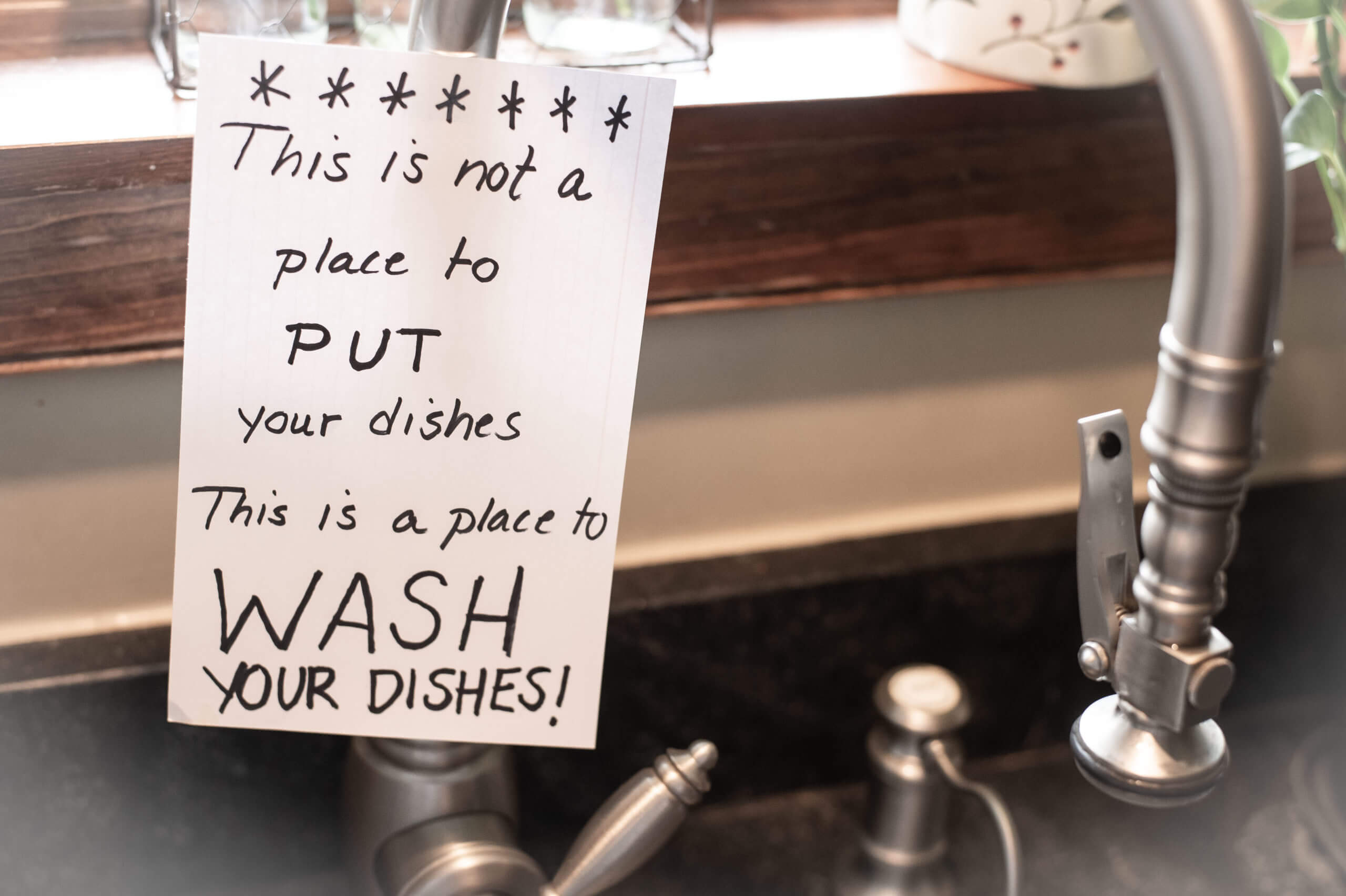 Sign taped to kitchen sink that reads, "This is not a place to PUT your dishes, this is a place to WASH YOUR DISHES"