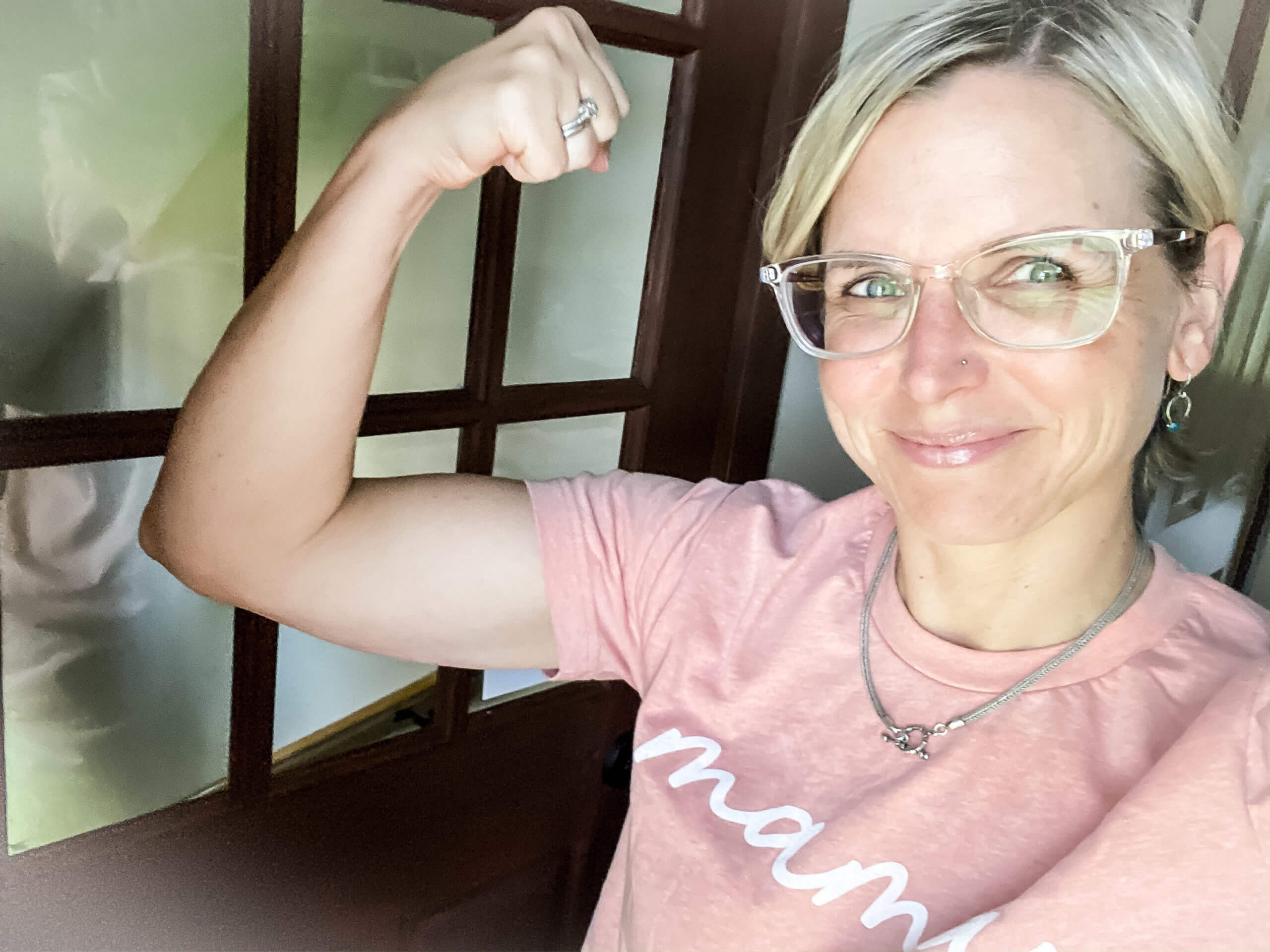 Mags, a white woman with short blond hair, blue eyes, and clear-rimmed glasses, takes a selfie with her other arm held up in a fist, making a muscle.