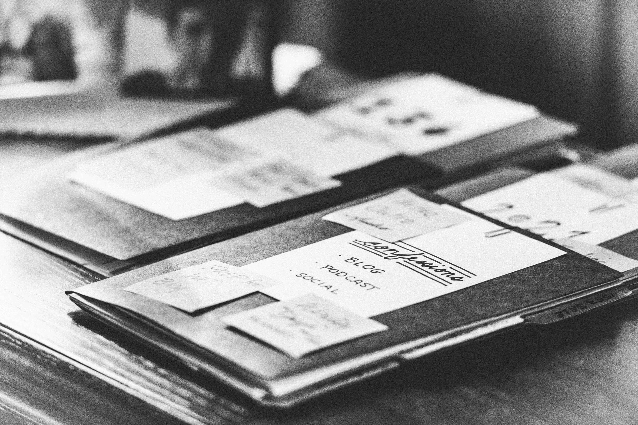 Black and white image of folders on a desk with 5x7 white note cards and post-it notes with writing on them.