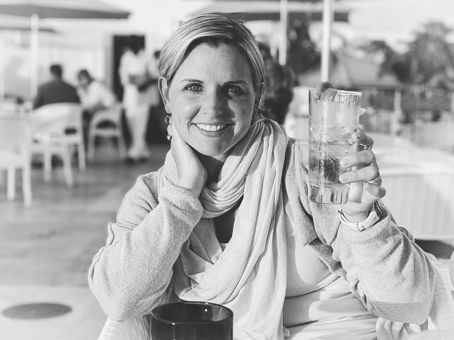 Black and white photo of a white woman with short blond hair sitting in a tropical setting outdoor restaurant. She's seated at a table holding up a drink in her left hand.
