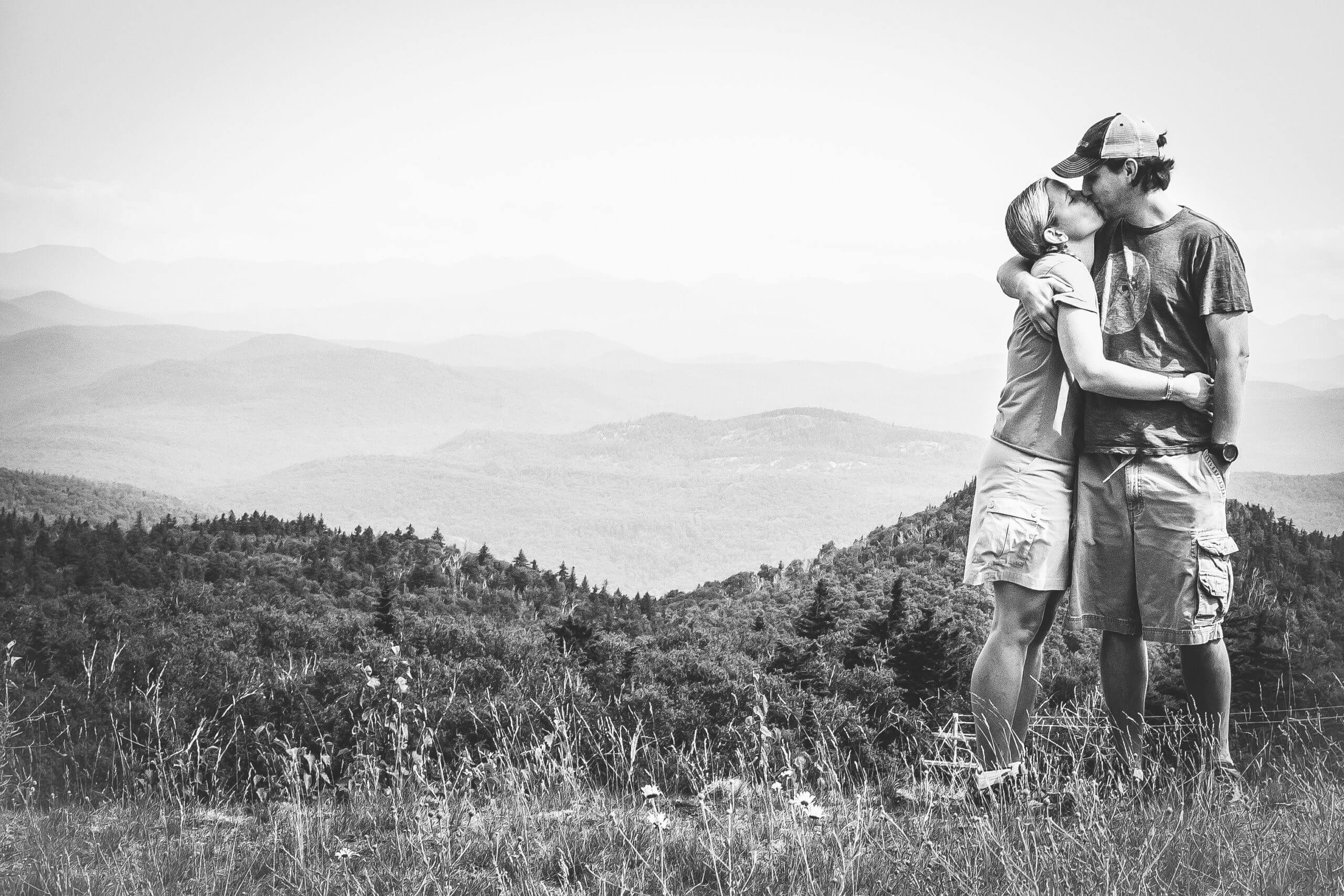 Black and white image of an Adirondack Mountain range with a white man and woman in summer clothes kissing in the forground on the right side of the photo.