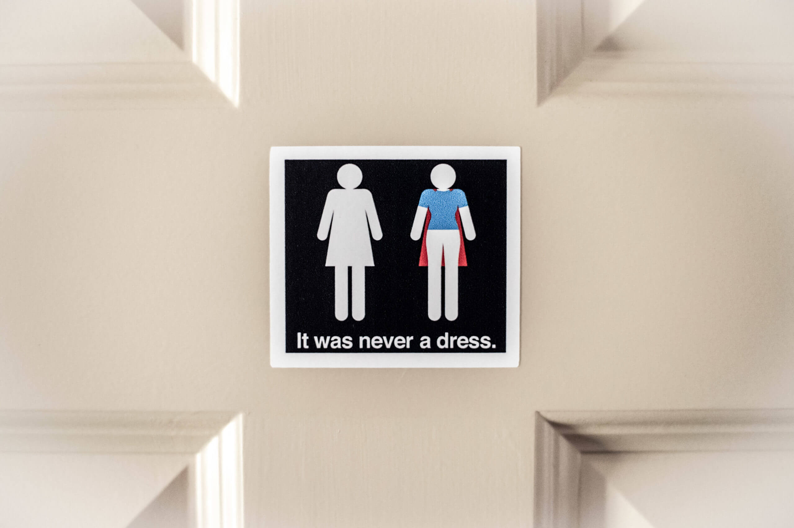 A color image of a 3 inch by 3 inch square sticker on a door that has the symbol for a woman's bathroom on the left in white and the same figure on the right with the outline of a cape. The sticker reads at the bottom, "It was never a dress."