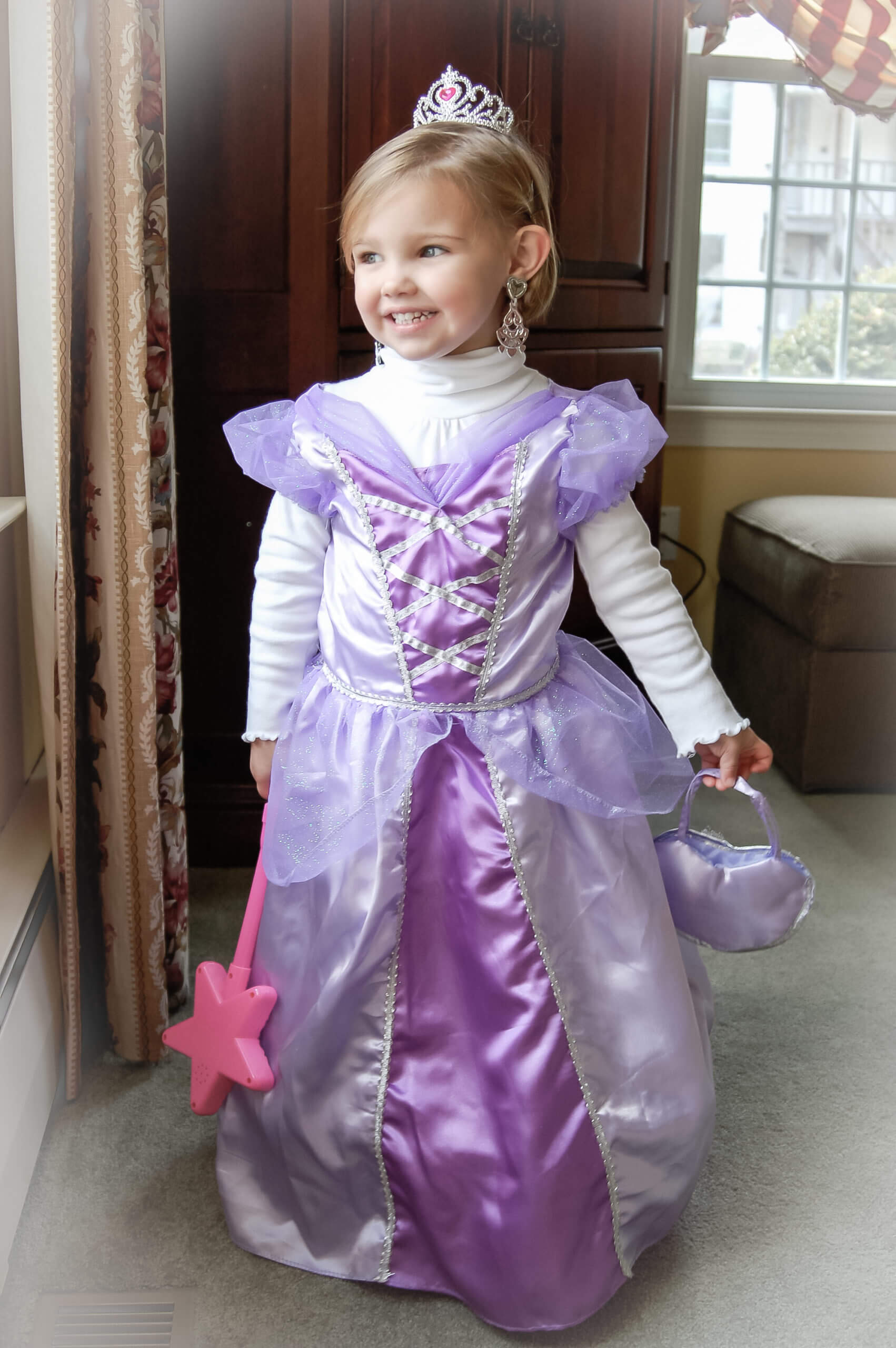 Color picture of three year old blog white girl in a purple princess costume, holding a pink plastic star wand in one hand, and a satin purple pocket book (that matches her dress) in her left hand.