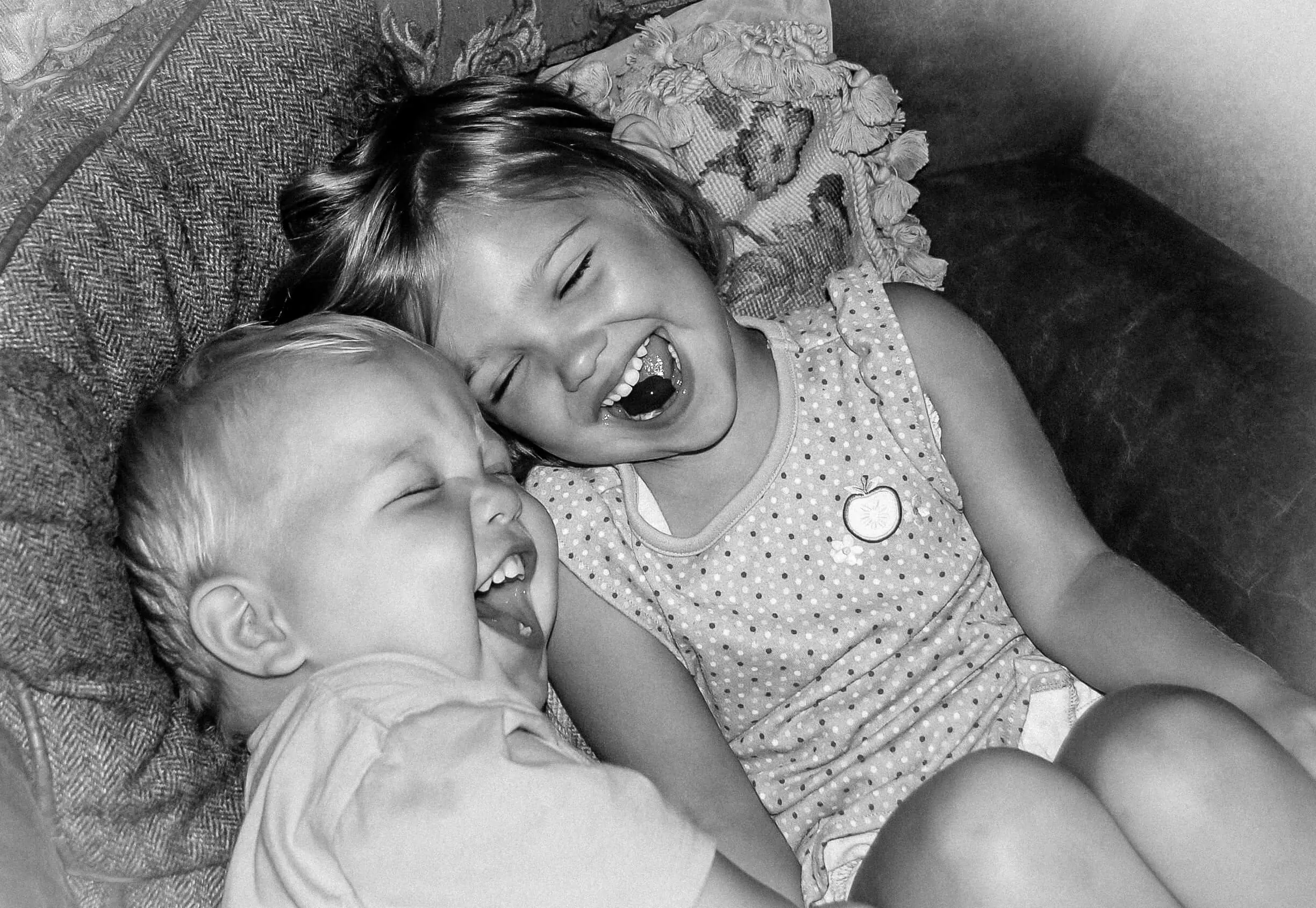 Black and white photo of two white children sitting in a chair giggling with their mouths open and their eyes closed.