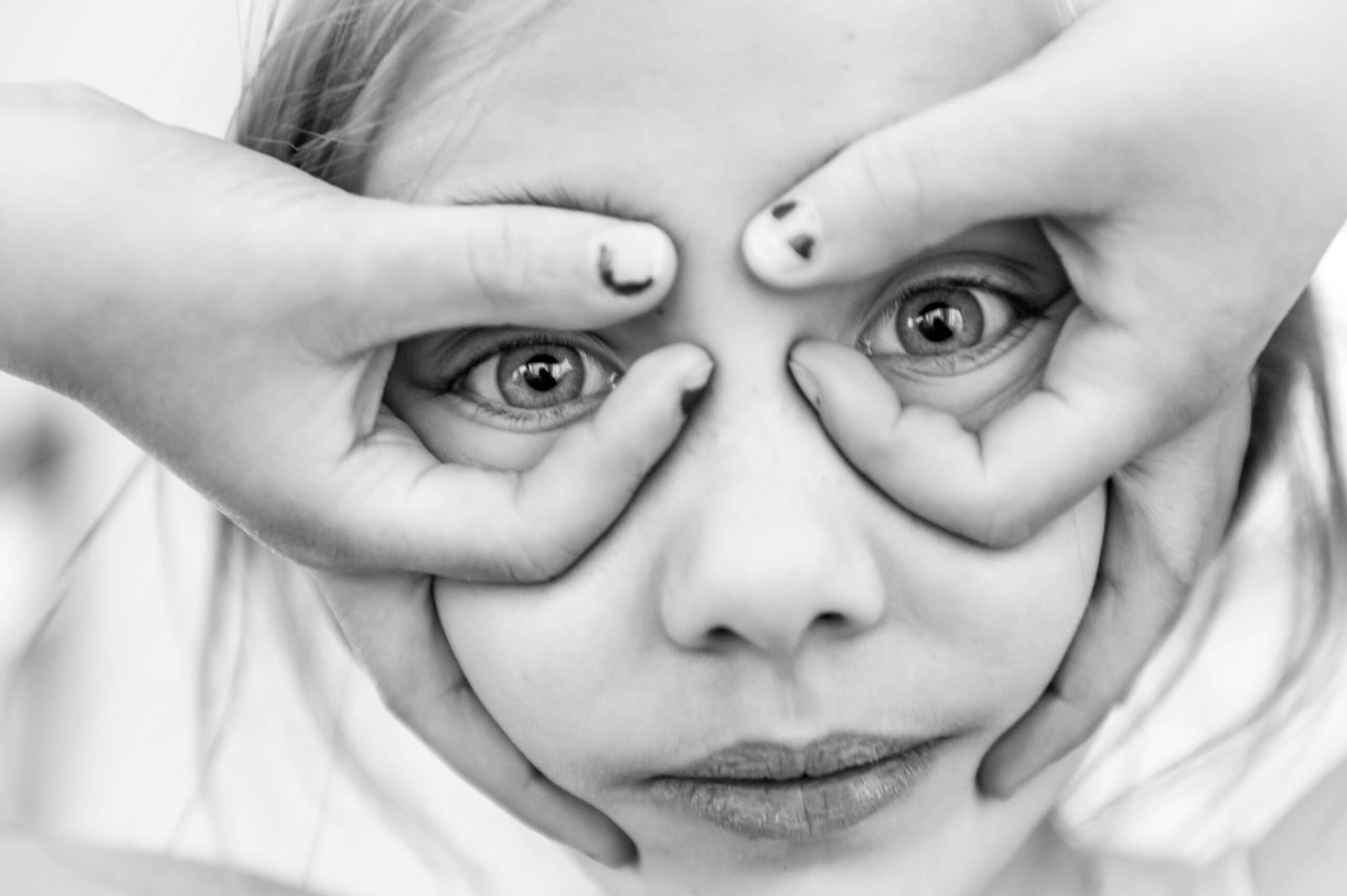 Black and white close-up of a white 8 year old girl with her hands upside down on her face and her pointer and thumb fingers making loops around her eyes.