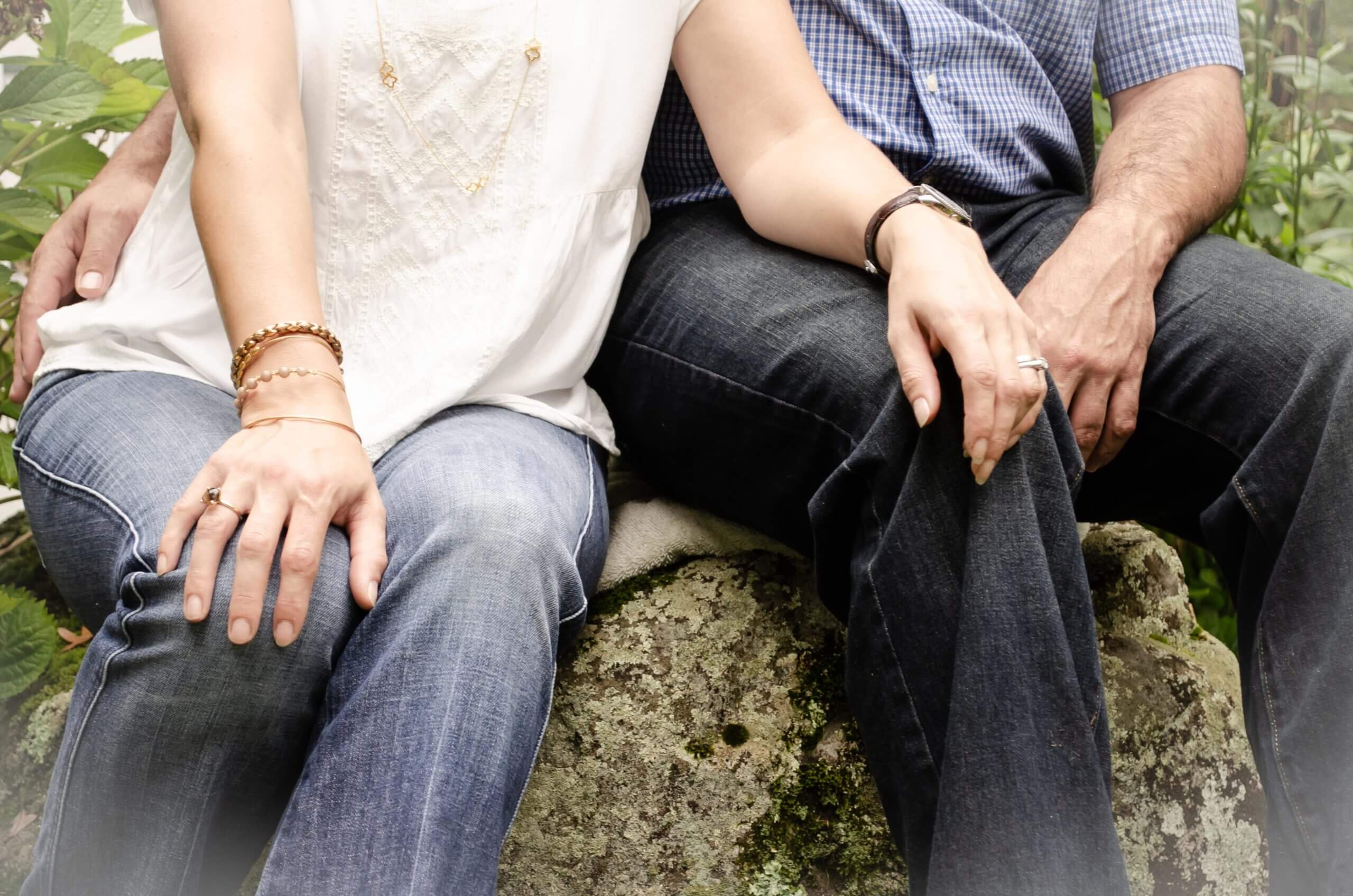 Close-up color photo of a white couple sitting next to one another on a rock outside. The woman's hand is on the man's thigh and his arm is wrapped around her waist. The photo is cut off at their shins and their torsos.